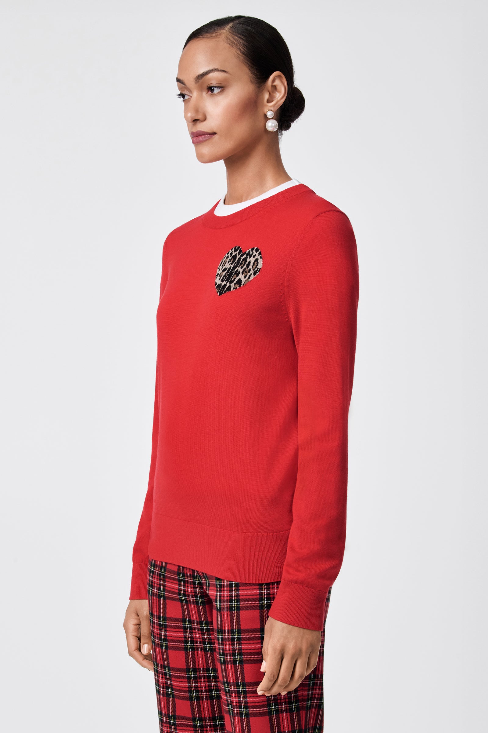 Woman wears the Jaguar Heart Crewneck Sweater in red, featuring a small leopard-print heart on the chest, paired with plaid pants and pearl earrings, standing against a simple light backdrop.