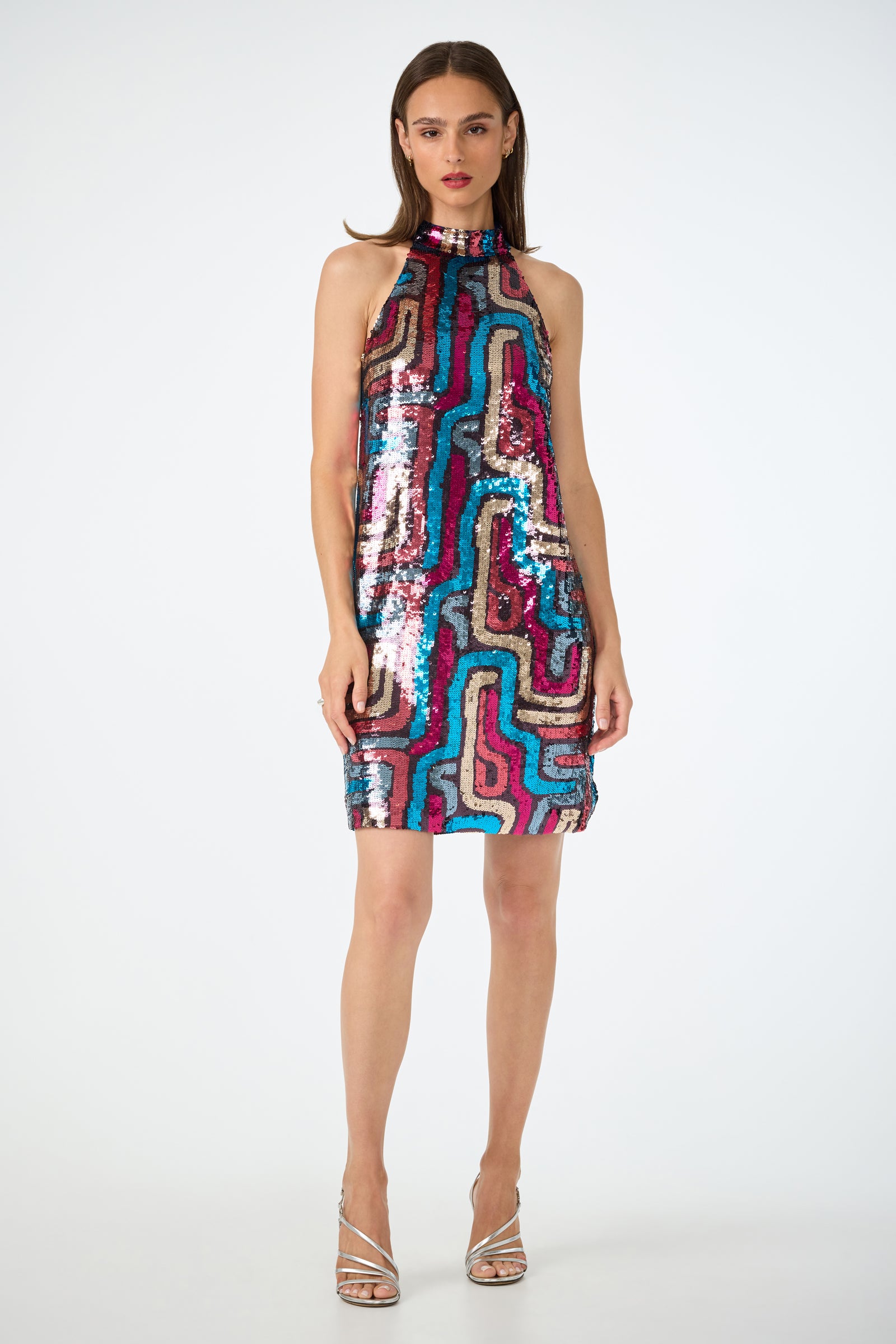 A woman faces forward in the Flamenco Sequin Mini Dress, a sleeveless, high-neck mini dress with colorful wavy sequins. She has straight brown hair, red lipstick, and wears silver strappy heels against a plain white background.