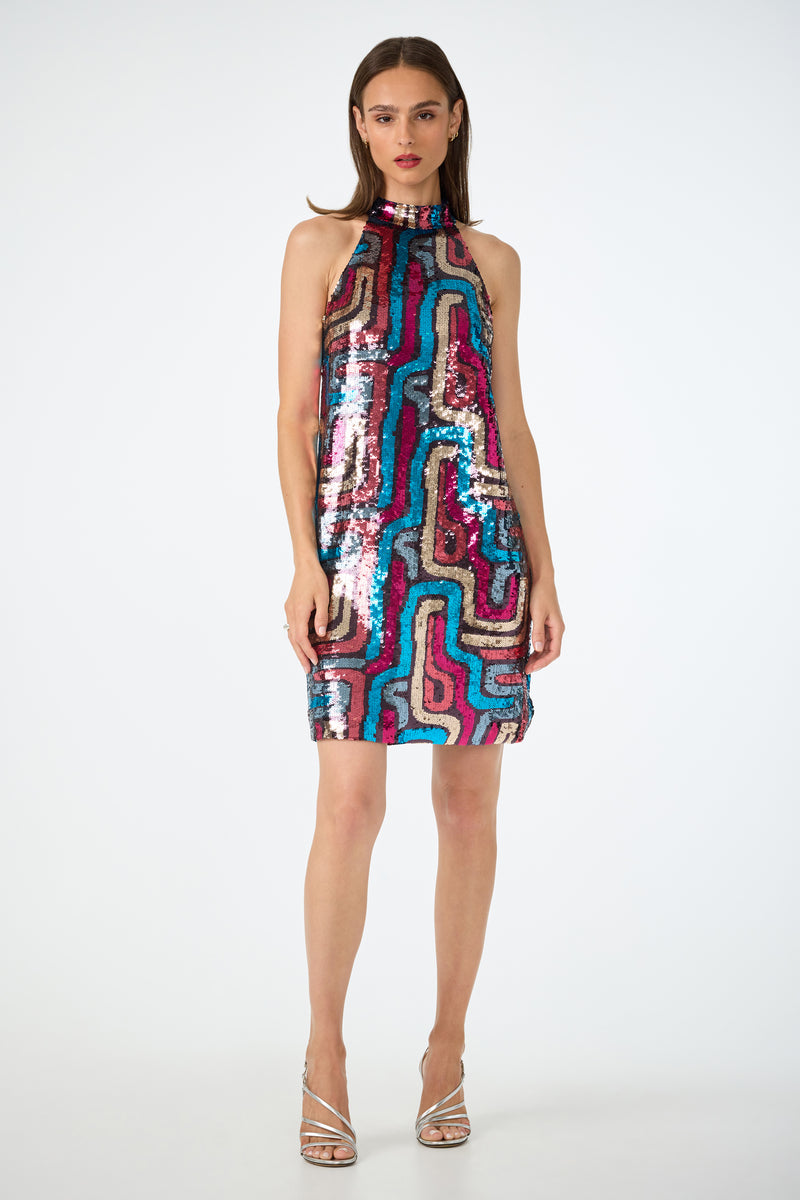 A woman faces forward in the Flamenco Sequin Mini Dress, a sleeveless, high-neck mini dress with colorful wavy sequins. She has straight brown hair, red lipstick, and wears silver strappy heels against a plain white background.