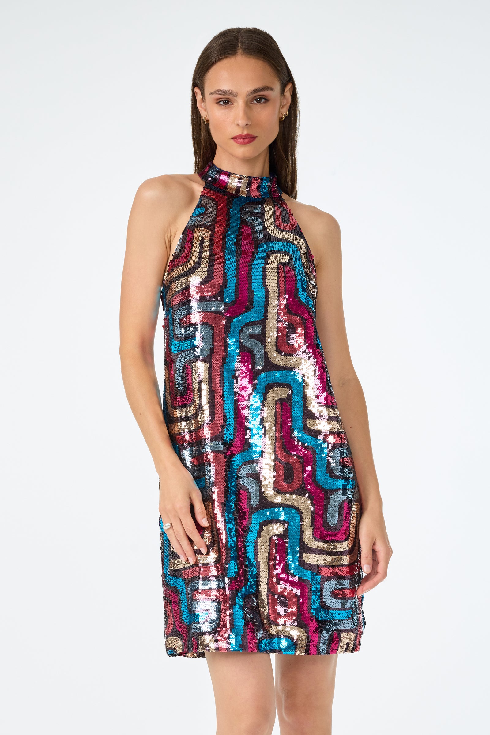 A woman wears the Flamenco Sequin Mini Dress—a sleeveless, high-neck, knee-length dress with vibrant blue, pink, gold, and black geometric sequins—standing against a plain white background.