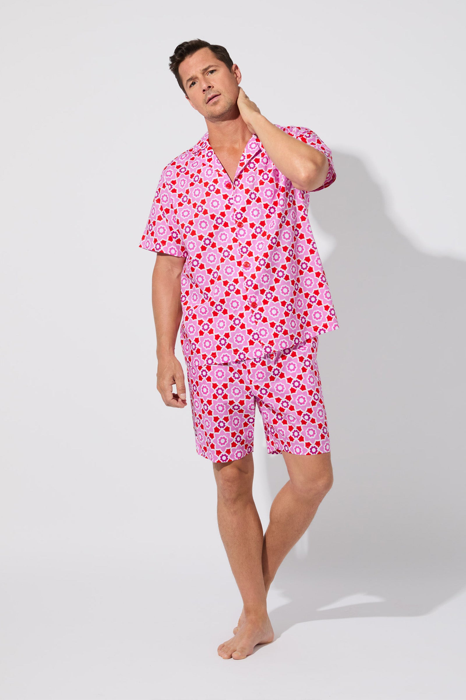 A man stands barefoot against a white background, wearing the Mosiac Tile Mens S/S Boxer Set— a matching short-sleeve button-up shirt and shorts with a bold pink and red geometric pattern. He looks relaxed, touching his neck with one hand.