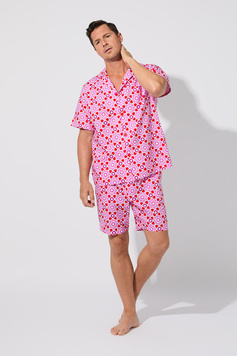 A man stands barefoot against a white background, wearing the Mosiac Tile Mens S/S Boxer Set— a matching short-sleeve button-up shirt and shorts with a bold pink and red geometric pattern. He looks relaxed, touching his neck with one hand.