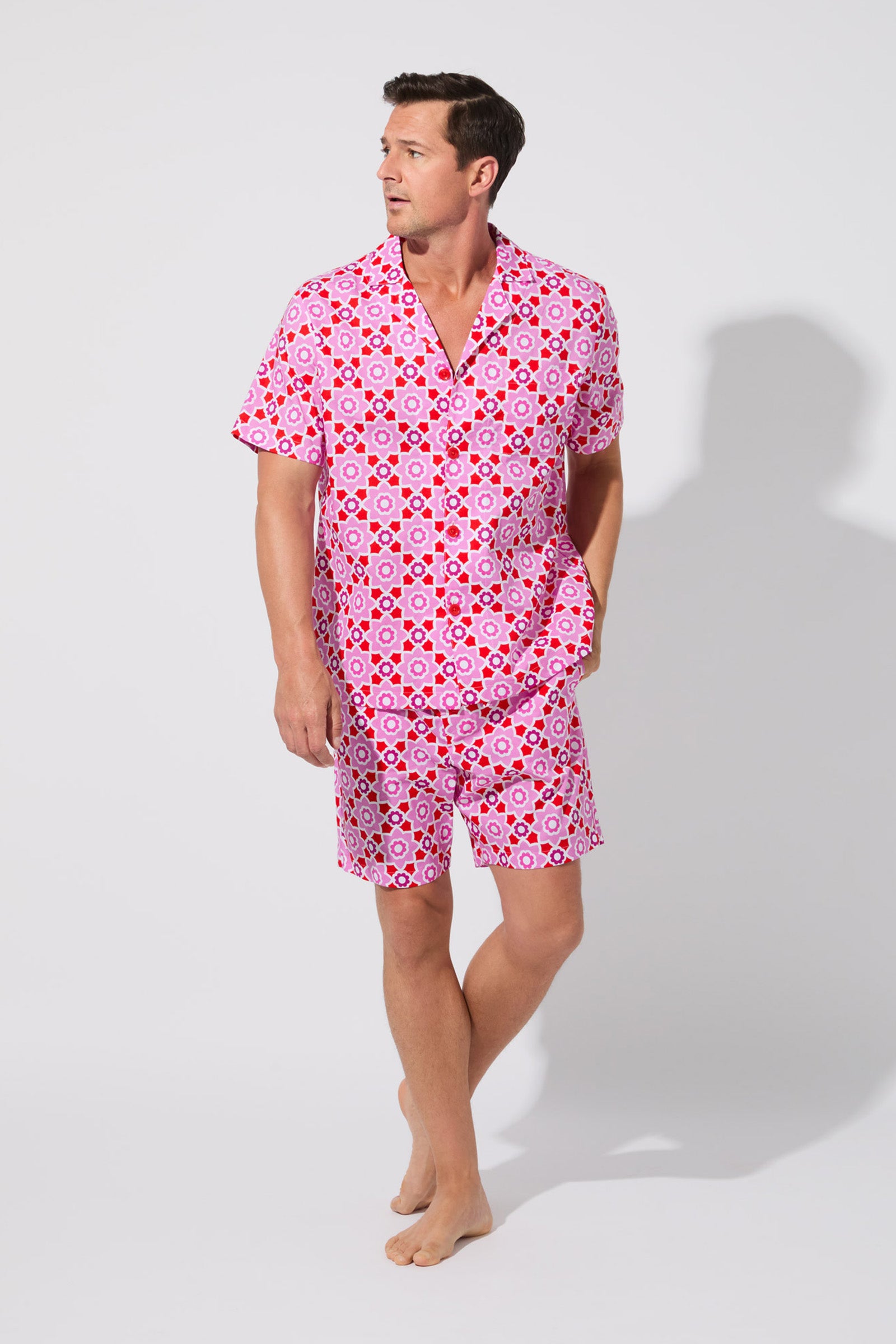 A man stands barefoot against a light background, wearing the Mosiac Tile Mens S/S Boxer Set—matching pink short-sleeve button-up shirt and shorts with a bold geometric pattern. He looks to his left, one hand in his pocket.