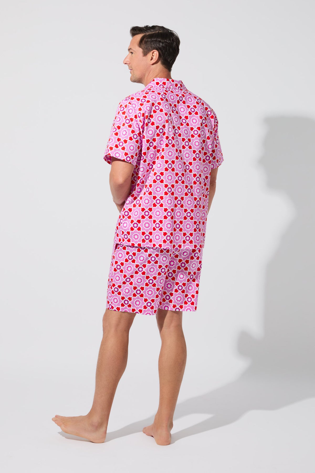 A barefoot man faces away, wearing the Mosiac Tile Mens S/S Boxer Set—matching pink shirt and shorts with a bold geometric circle and diamond pattern—against a plain, light-colored background.