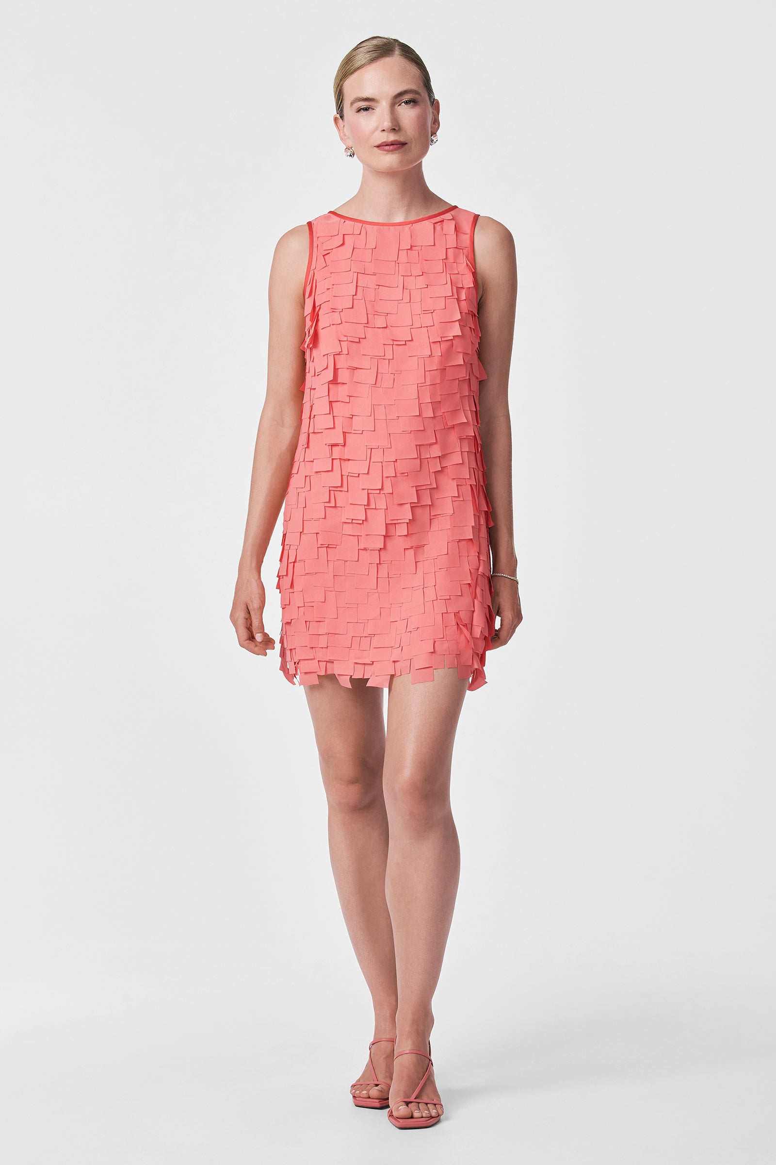 A woman poses against a white background in the Valdis Sleeveless Shift Dress - Flutter Chiffon, a coral pink mini dress with layered rectangular details, styled with matching strappy heels and her hair pulled back.