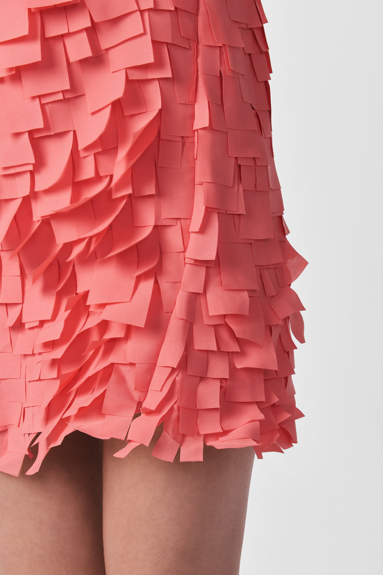 A close-up of the Valdis Sleeveless Shift Dress - Flutter Chiffon in coral pink, showcasing layered rectangular chiffon pieces for a textured, 3D look. Only part of the wearer's legs is visible.
