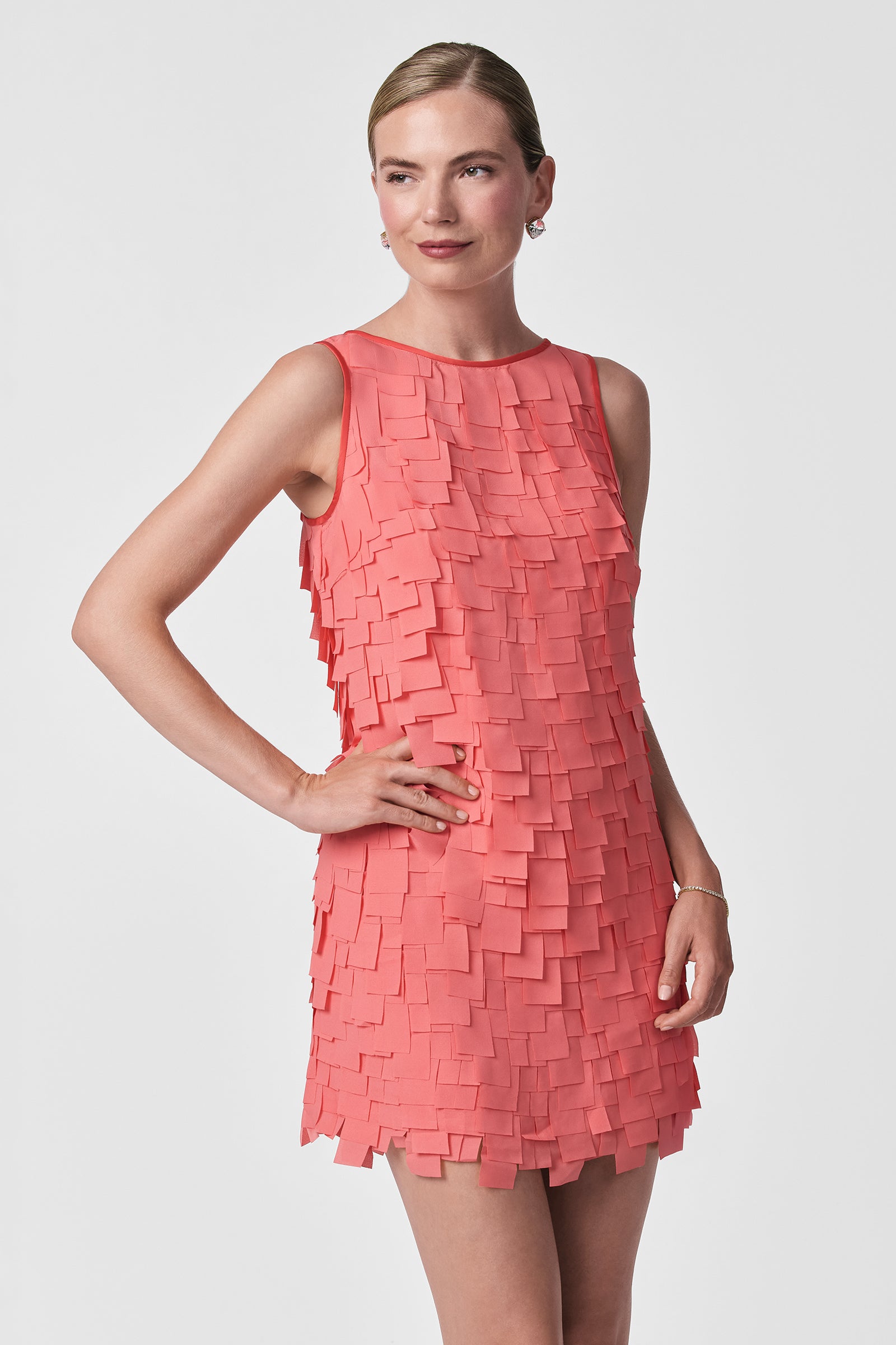 A woman stands against a plain background wearing the Valdis Sleeveless Shift Dress - Flutter Chiffon in coral, featuring square, layered fabric pieces. Her hair is pulled back, one hand on her hip, and she smiles slightly.