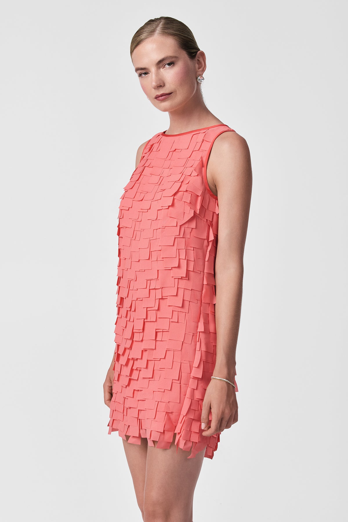 A woman wears the Valdis Sleeveless Shift Dress - Flutter Chiffon, featuring a coral-pink hue and layered square fabric details, as she stands against a plain white background, looking slightly to the side.