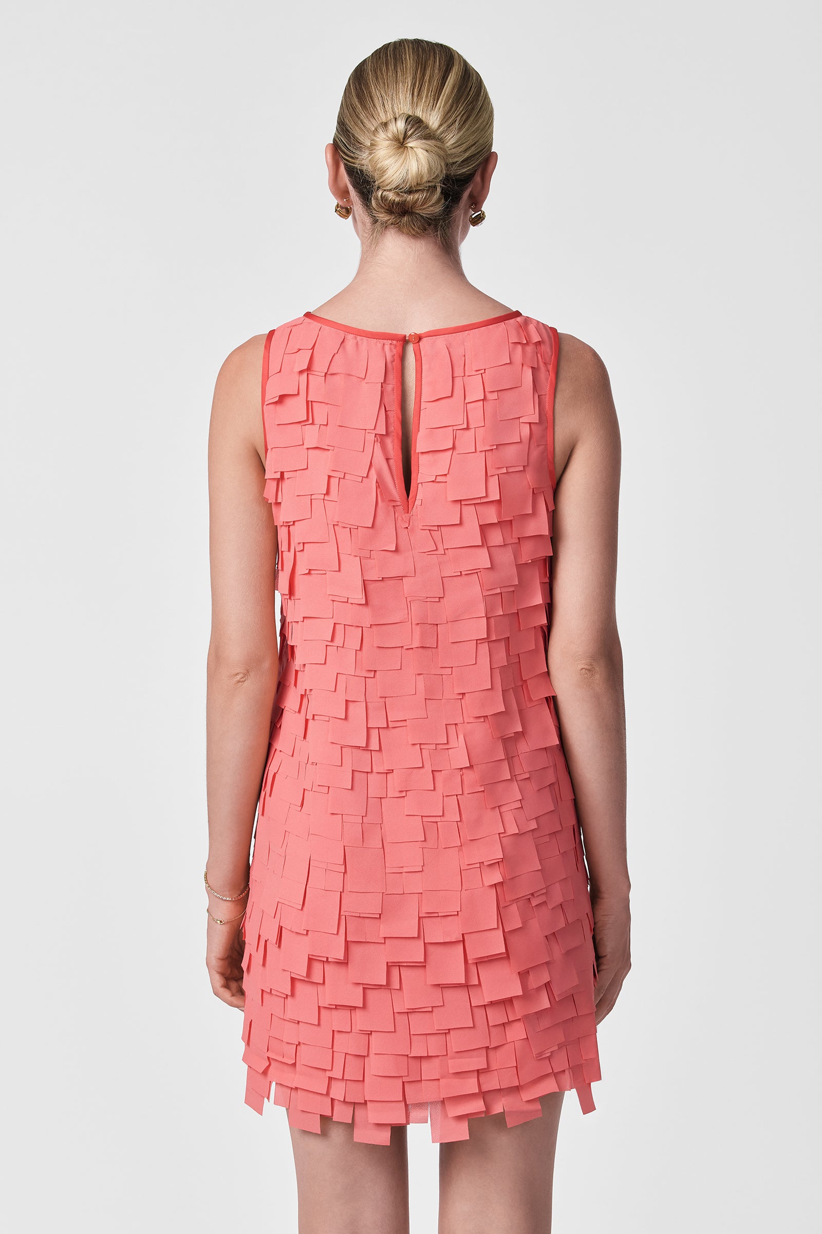 A woman with blonde hair in a neat bun is shown from the back, modeling the Valdis Sleeveless Shift Dress - Flutter Chiffon in coral, featuring layered square chiffon pieces, against a plain white background.