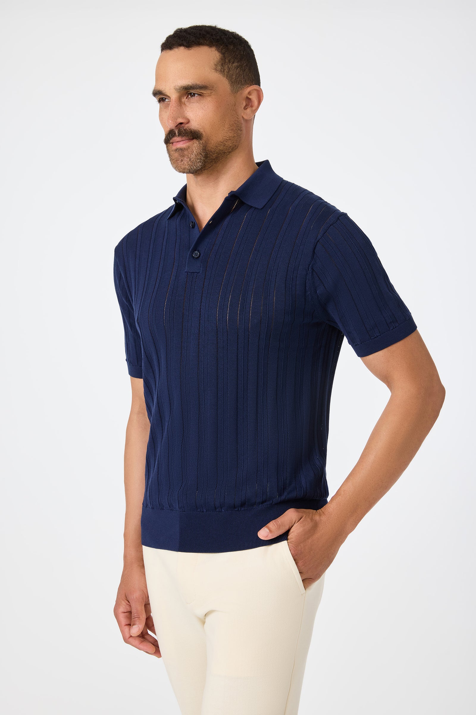 A man with short dark hair and a beard wears the Kensington Short-Sleeve Polo in navy blue and cream-colored pants, posing with one hand in his pocket against a plain white background.