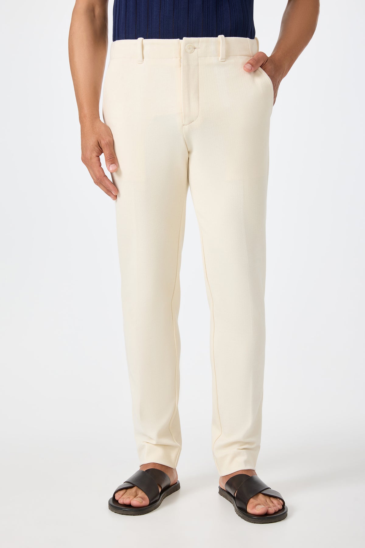 Someone wearing the Alex Slim Trouser in ecru, paired with a navy top and black slide sandals, stands with one hand in their pocket against a plain white background.