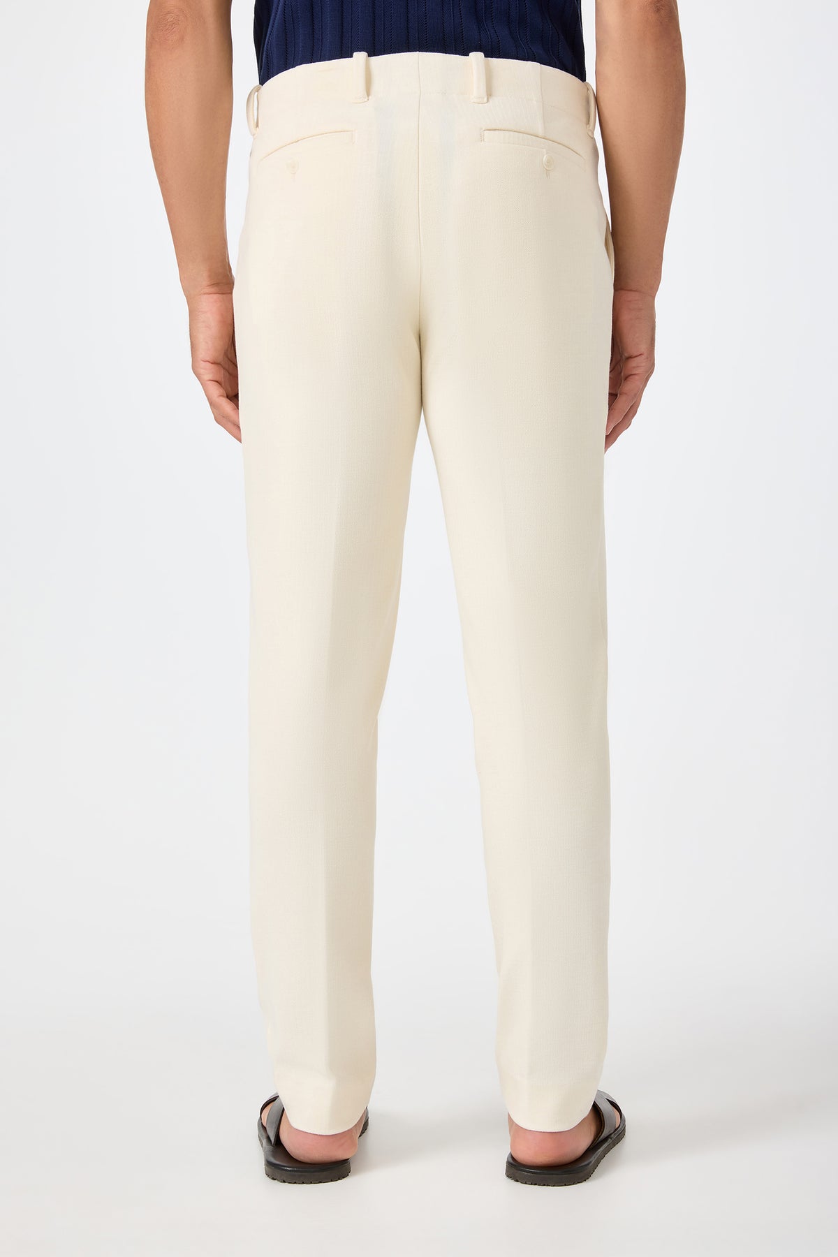 A person is seen from behind wearing the Alex Slim Trouser in Ecru with a dark shirt and black sandals, standing against a plain white background.