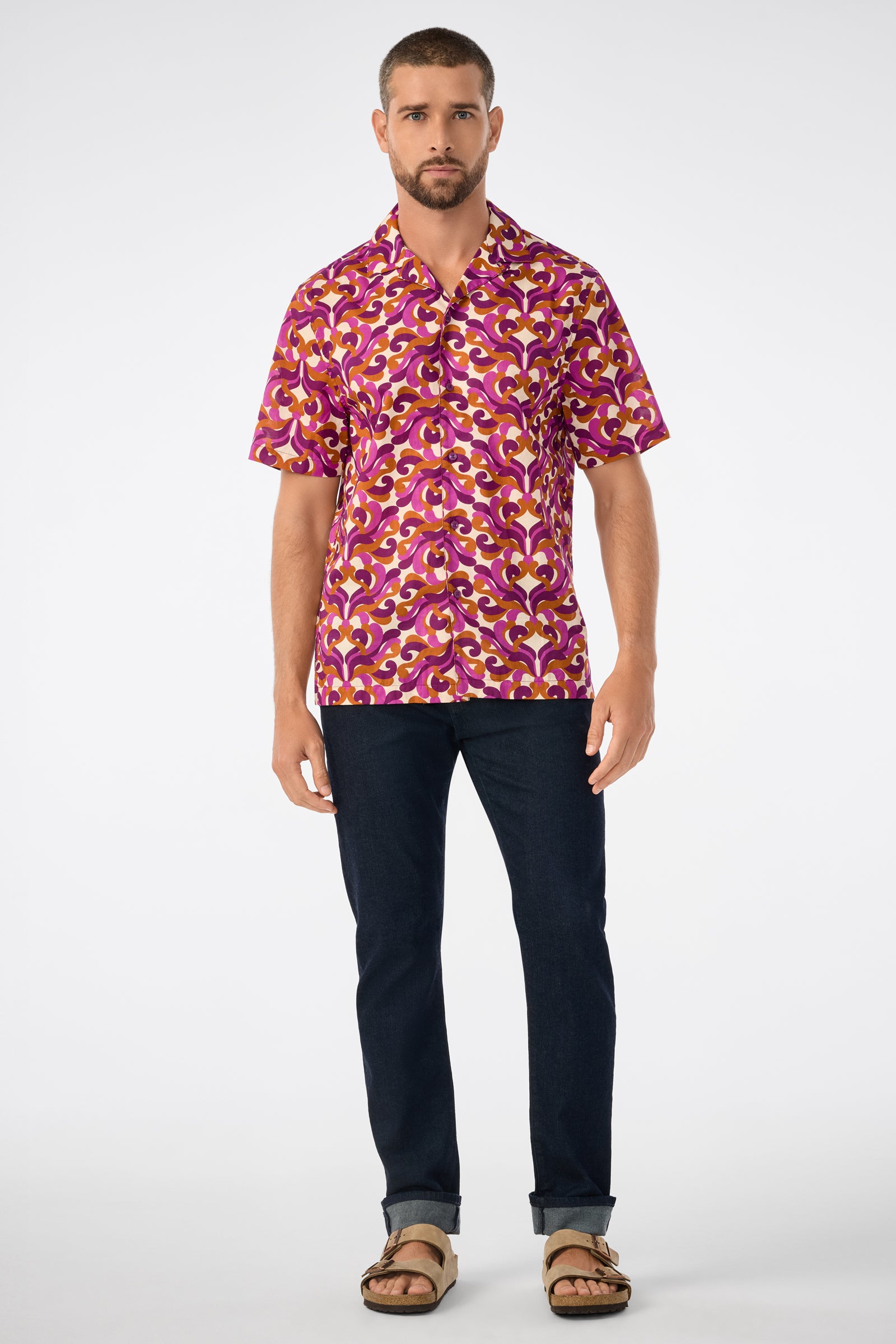 A man with short brown hair and a beard wears the Felix Ss Camp Shirt, featuring a bright pink, yellow, and white pattern, paired with dark jeans and brown sandals, standing against a plain white background.