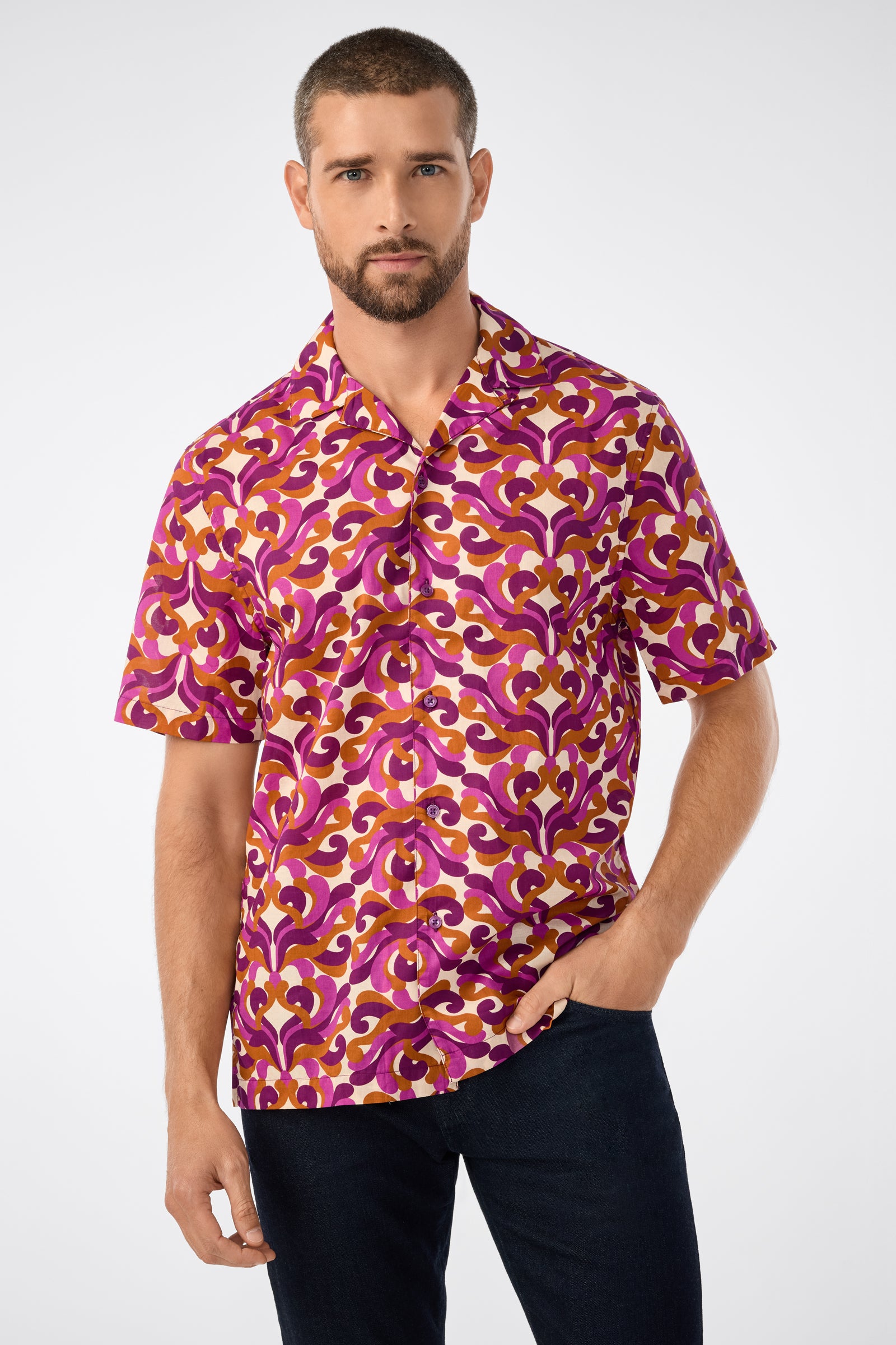 A man with short hair and a beard wears the Felix Ss Camp Shirt, a bold short-sleeve button-up featuring a vibrant purple, pink, and yellow geometric pattern. He stands with one hand in his pocket against a plain background.