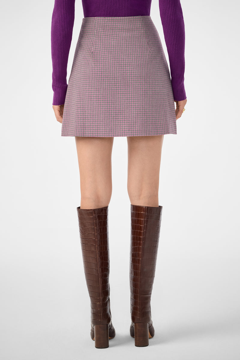A person stands facing away, wearing a purple long-sleeve top, the High Rise Rico Skirt (a plaid A-line mini), and tall brown heeled boots against a plain, light-colored background.