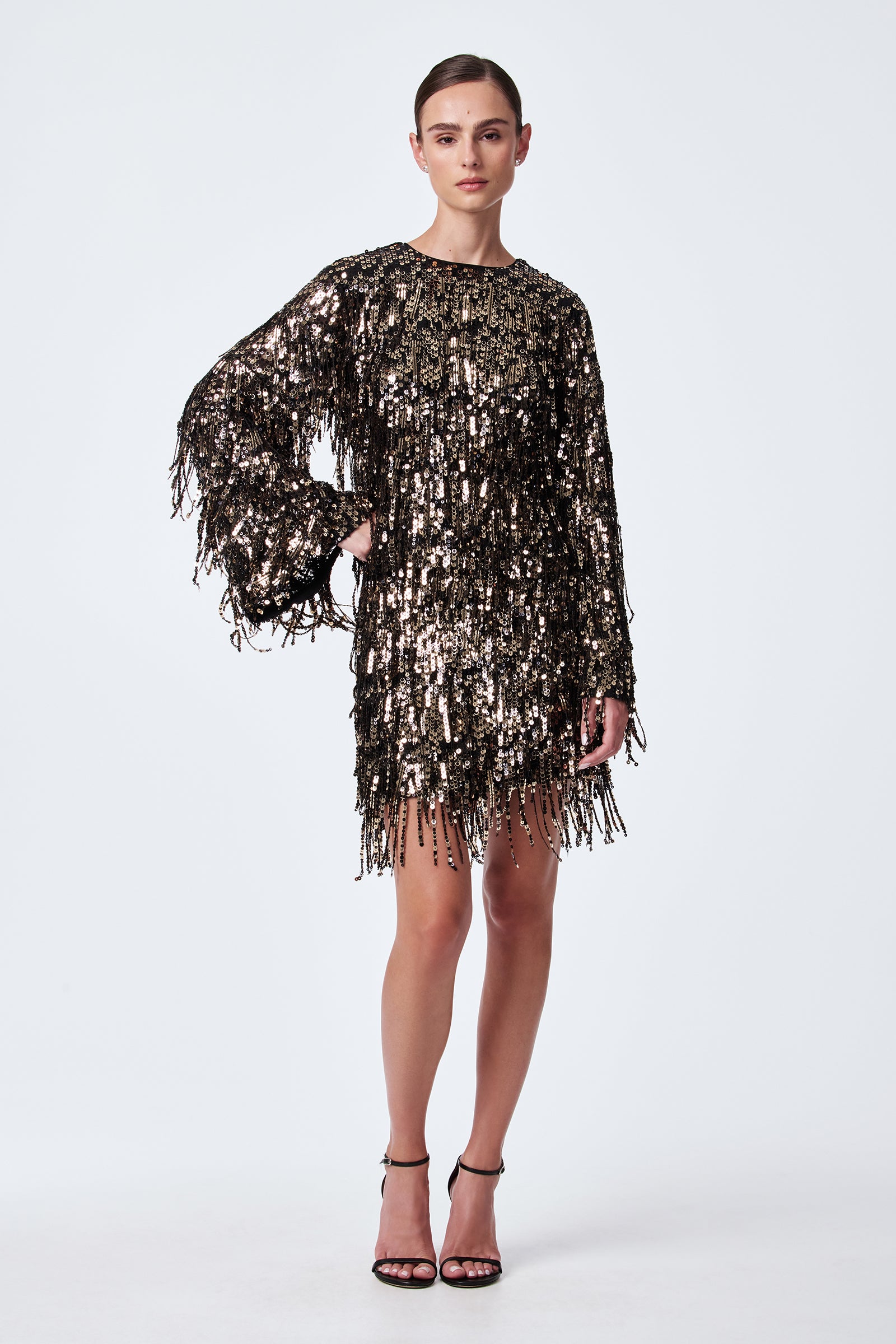 A woman poses against a plain background in the Satine Sequin Fringe Top with long sleeves, paired with a knee-length skirt and black high-heeled sandals. Her hair is slicked back for a sleek look.