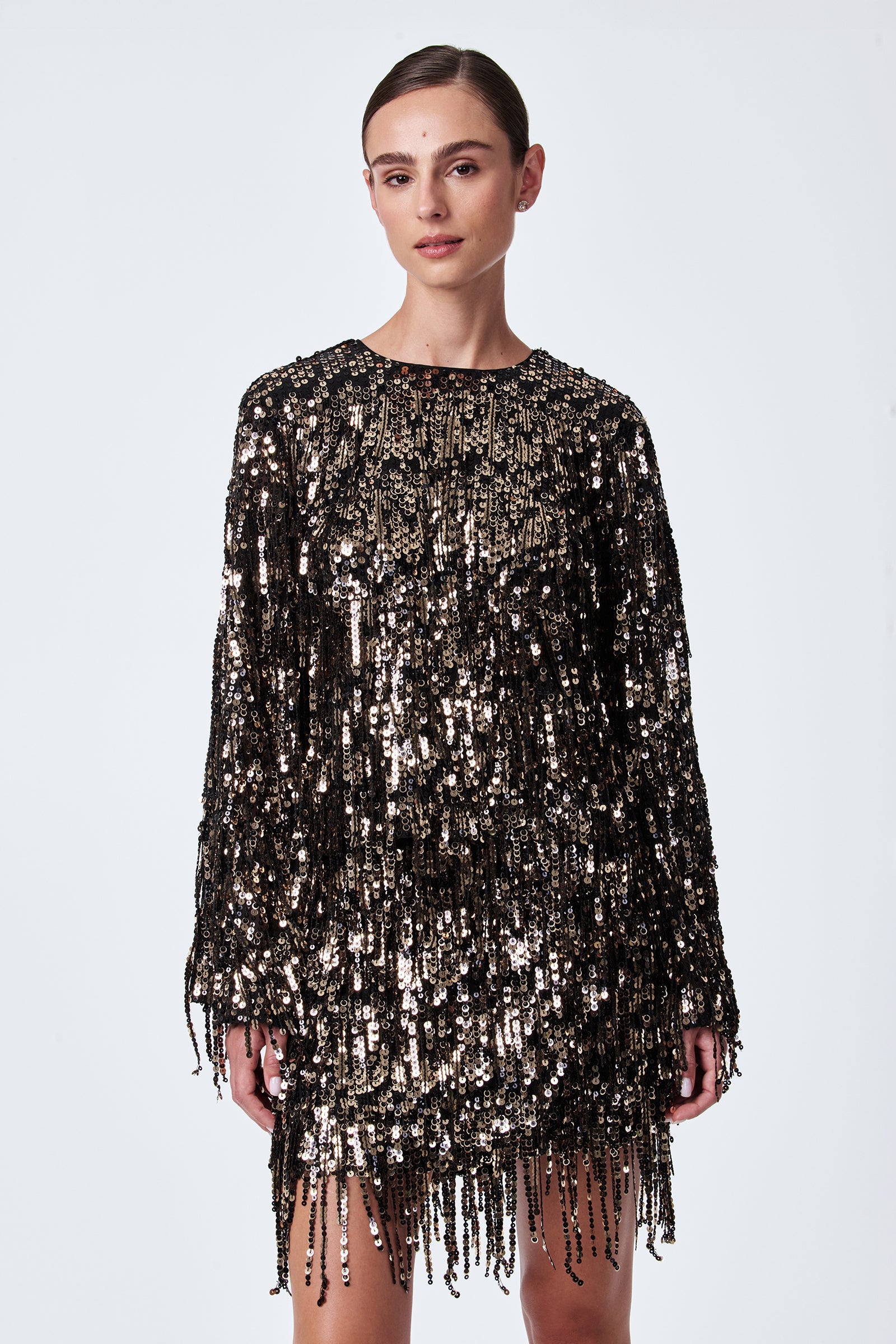 A woman stands against a plain background wearing the Satine Sequin Fringe Top, which features black and gold shimmering sequins with fringe detailing for a sparkling, textured effect.
