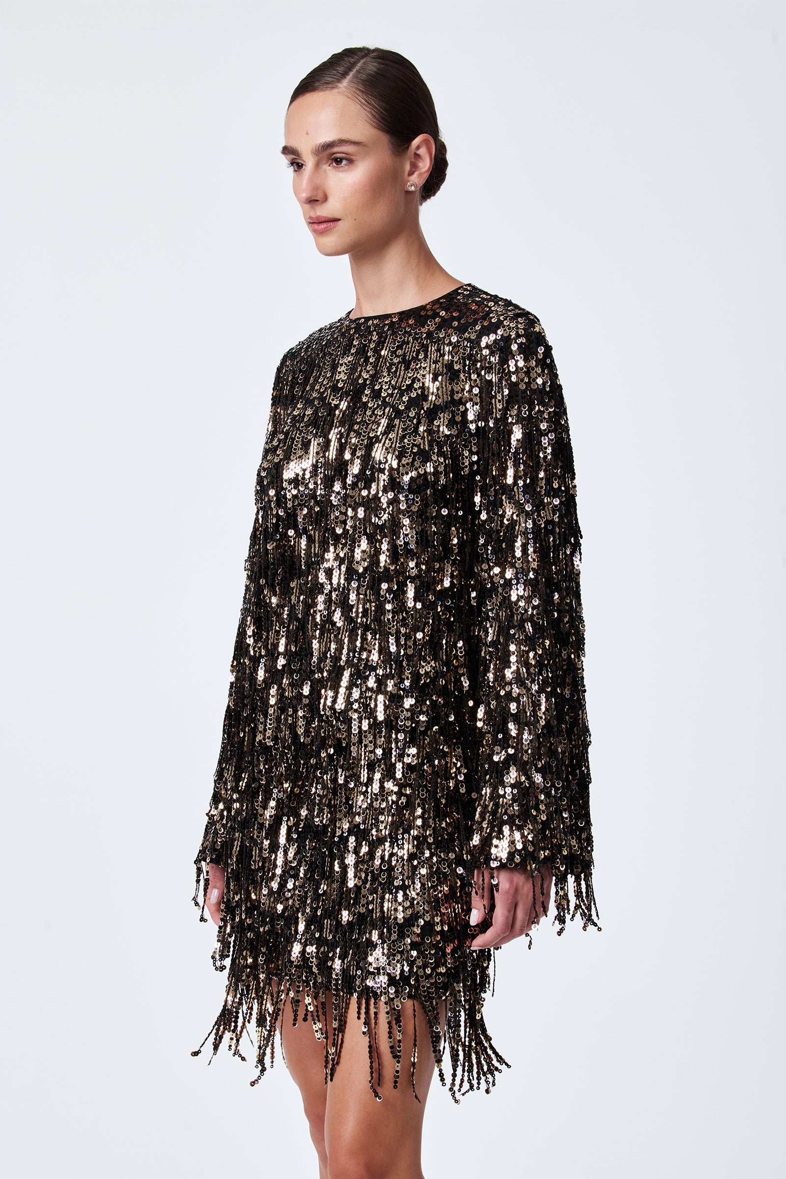 A woman stands against a plain background, wearing the Satine Sequin Fringe Top in black with shiny gold and black sequins and fringe details, her hair styled in a sleek low bun.