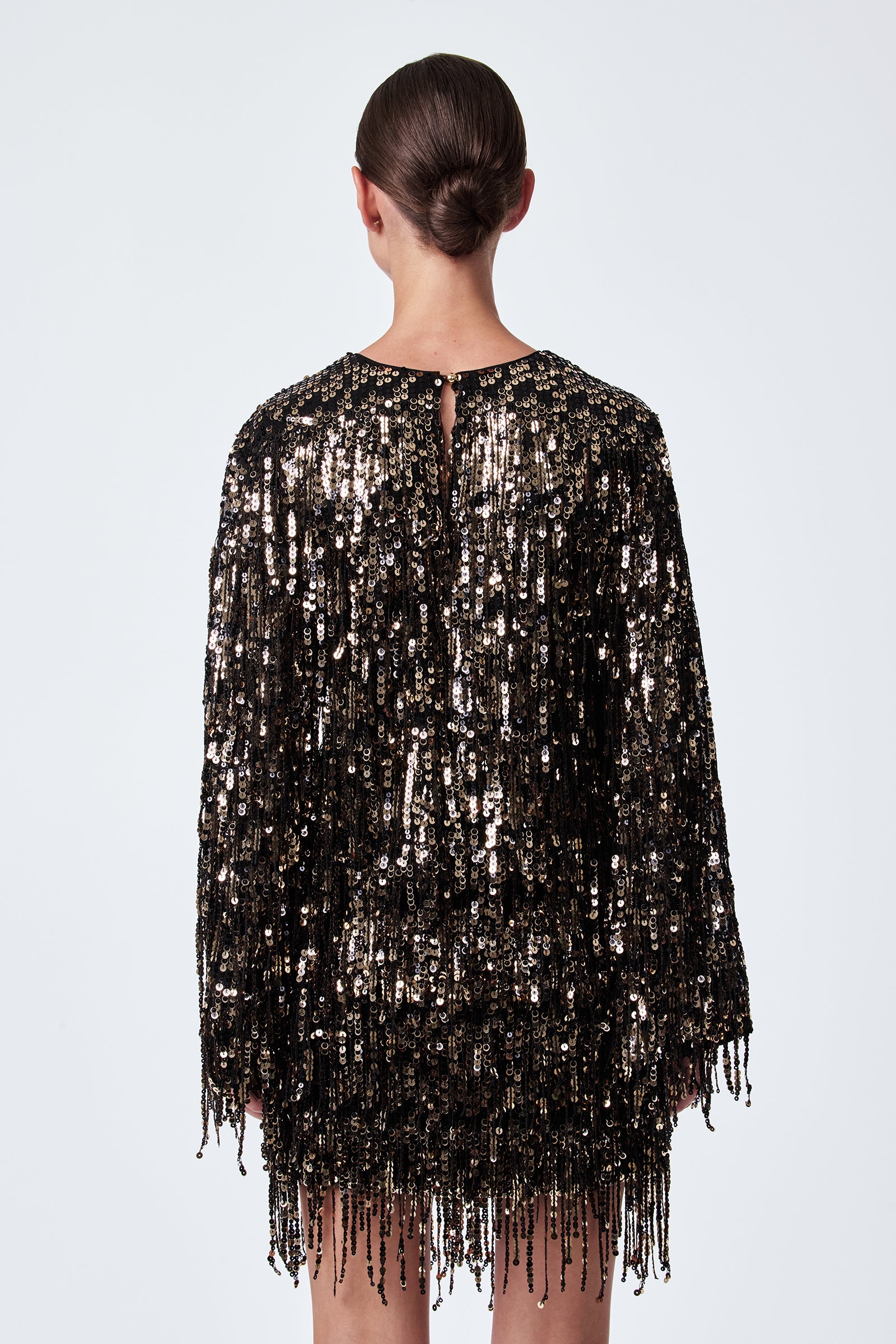 A person with dark hair in a low bun is shown from behind, wearing the Satine Sequin Fringe Top—black, long-sleeved, and adorned with shimmering gold and black sequins and fringe—against a plain white background.