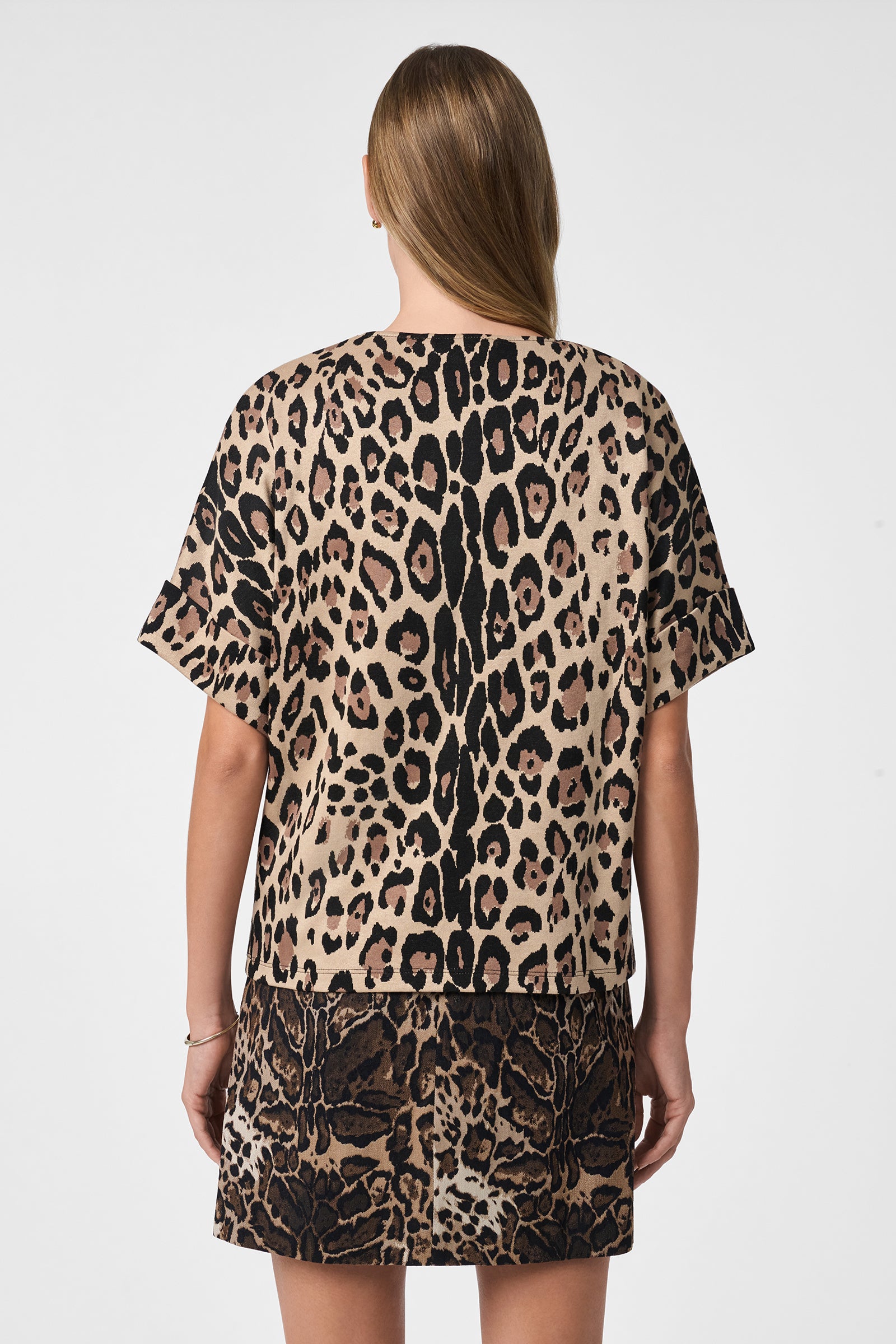 A woman with straight light brown hair faces away, wearing the loose-fitting Stagehands Top in leopard print and a matching skirt, set against a plain white background.