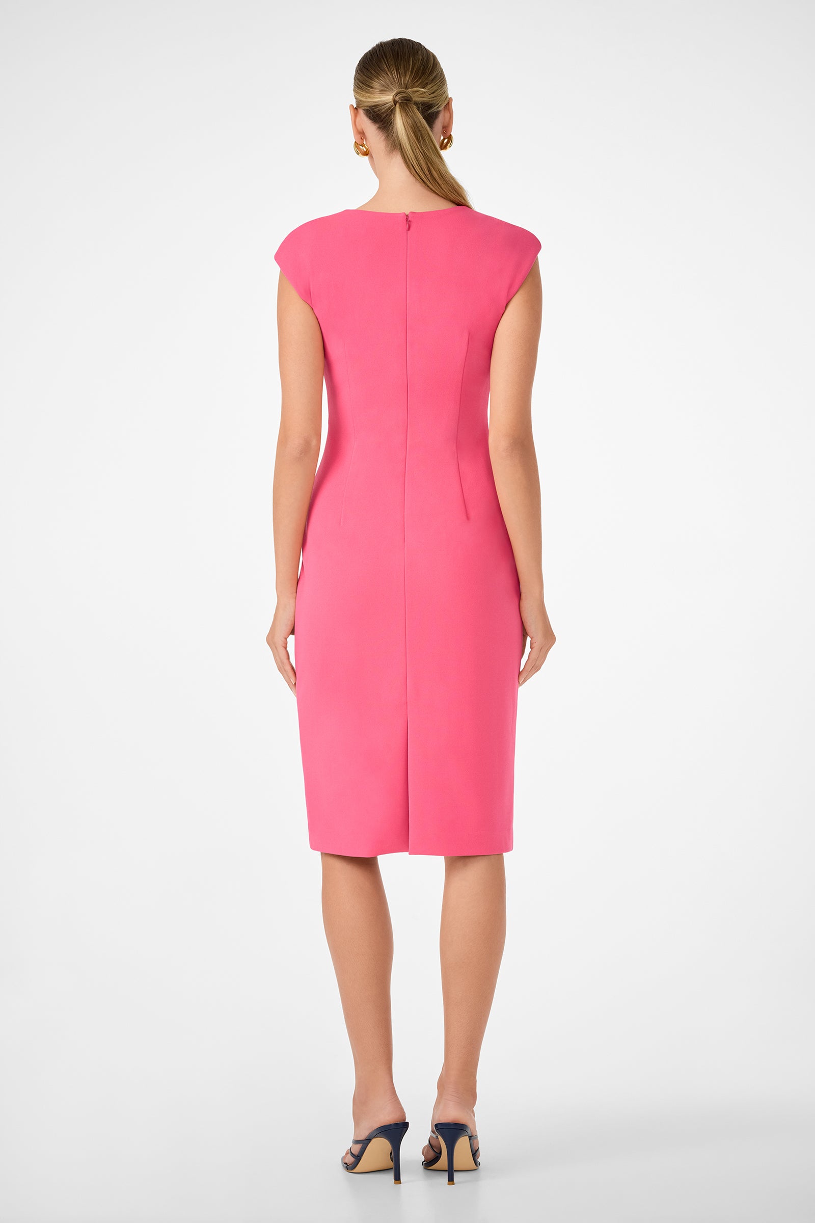 A woman faces away, wearing the Vibrant Dress—a fitted, knee-length, sleeveless pink dress—with black heeled sandals. Her hair is neatly pulled back against a plain white background.