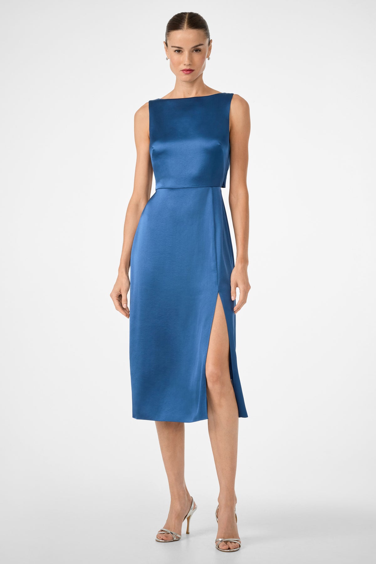 A woman wears the Mochi Sleeveless Midi Dress in Sumi Blue, featuring a fitted silhouette and high side slit, styled with silver high-heeled sandals. She stands against a plain white background.