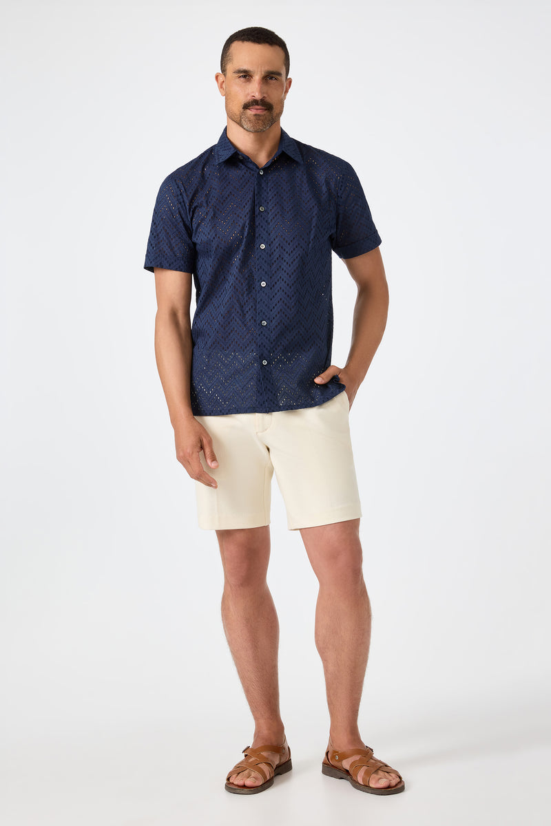 A man stands against a plain backdrop wearing the Branson Shirt - Cotton Eyelet in navy, paired with cream shorts and brown sandals. One hand is in his pocket and he has a neutral expression.