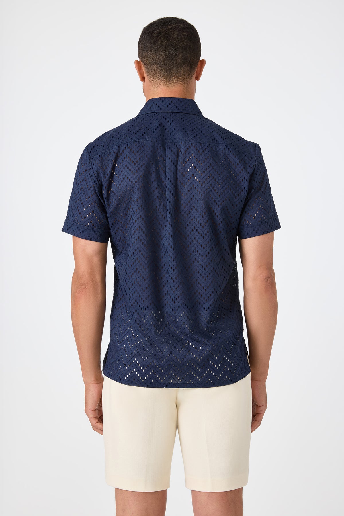 A man faces away, wearing the Branson Shirt - Cotton Eyelet in navy with cream-colored shorts, set against a plain white background.