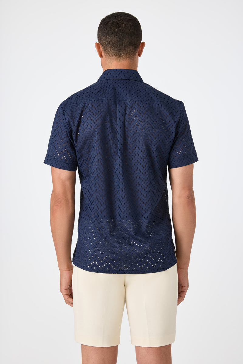 A man faces away, wearing the Branson Shirt - Cotton Eyelet in navy with cream-colored shorts, set against a plain white background.