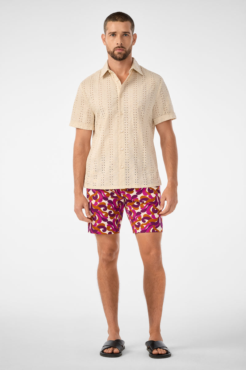 A man with short hair and a trimmed beard models the Branson Shirt—a beige short-sleeve button-up with eyelet details—pairing it with patterned pink and yellow shorts and black sandals against a plain background.