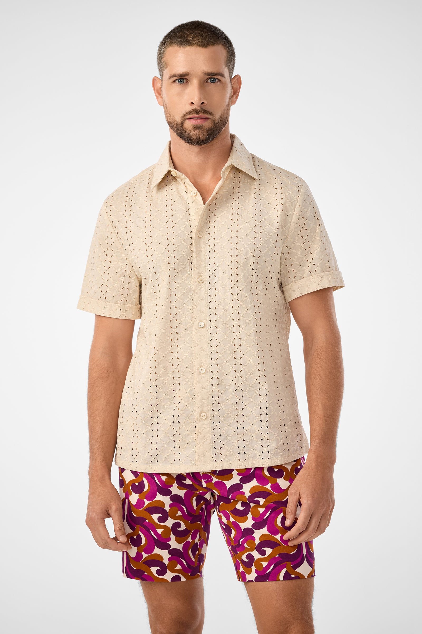 A man with short hair and a beard wears the Branson Shirt—a beige, short-sleeved, button-up shirt with an eyelet pattern—paired with vibrant purple and orange patterned shorts, standing against a plain white background.