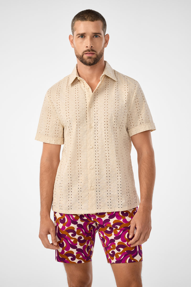 A man with short hair and a beard wears the Branson Shirt—a beige, short-sleeved, button-up shirt with an eyelet pattern—paired with vibrant purple and orange patterned shorts, standing against a plain white background.