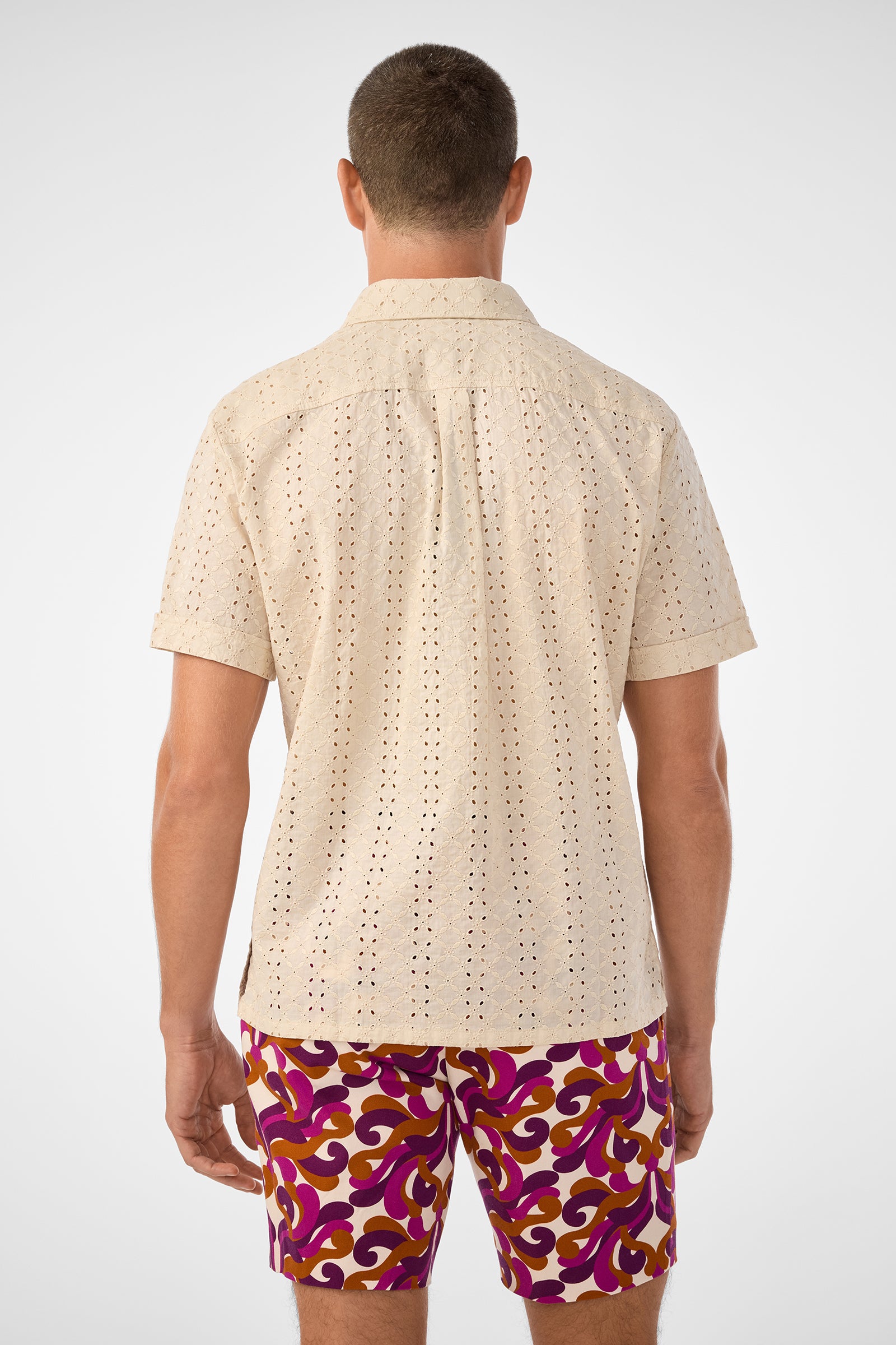 A man is seen from the back in a short-sleeved, beige Branson Shirt and vibrant purple, yellow, and red patterned shorts against a plain light background.