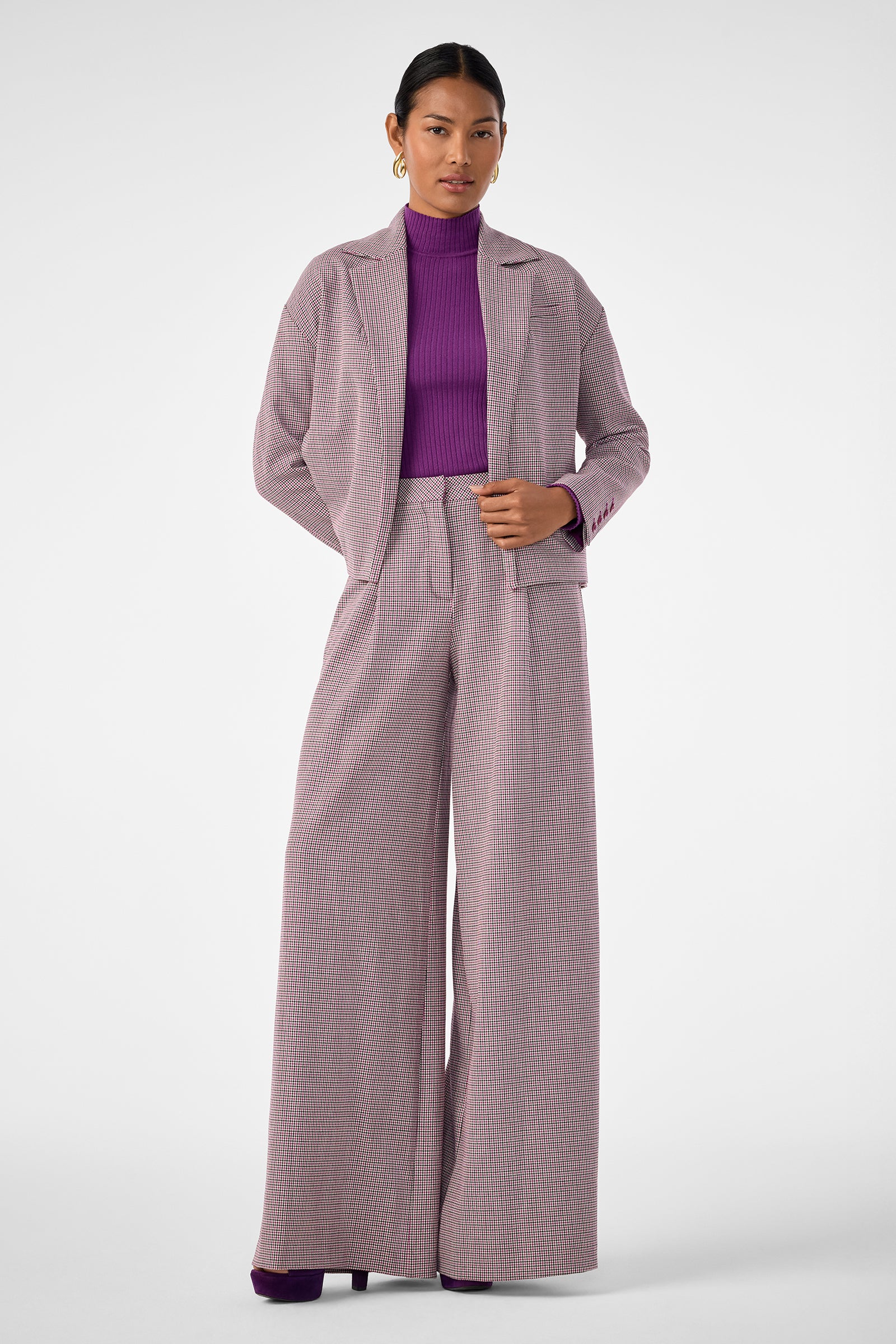 A woman stands facing forward against a plain, light background, wearing the Ayla Jacket—a loose-fitting plaid blazer paired with wide-leg pants over a purple turtleneck. Her hair is pulled back.