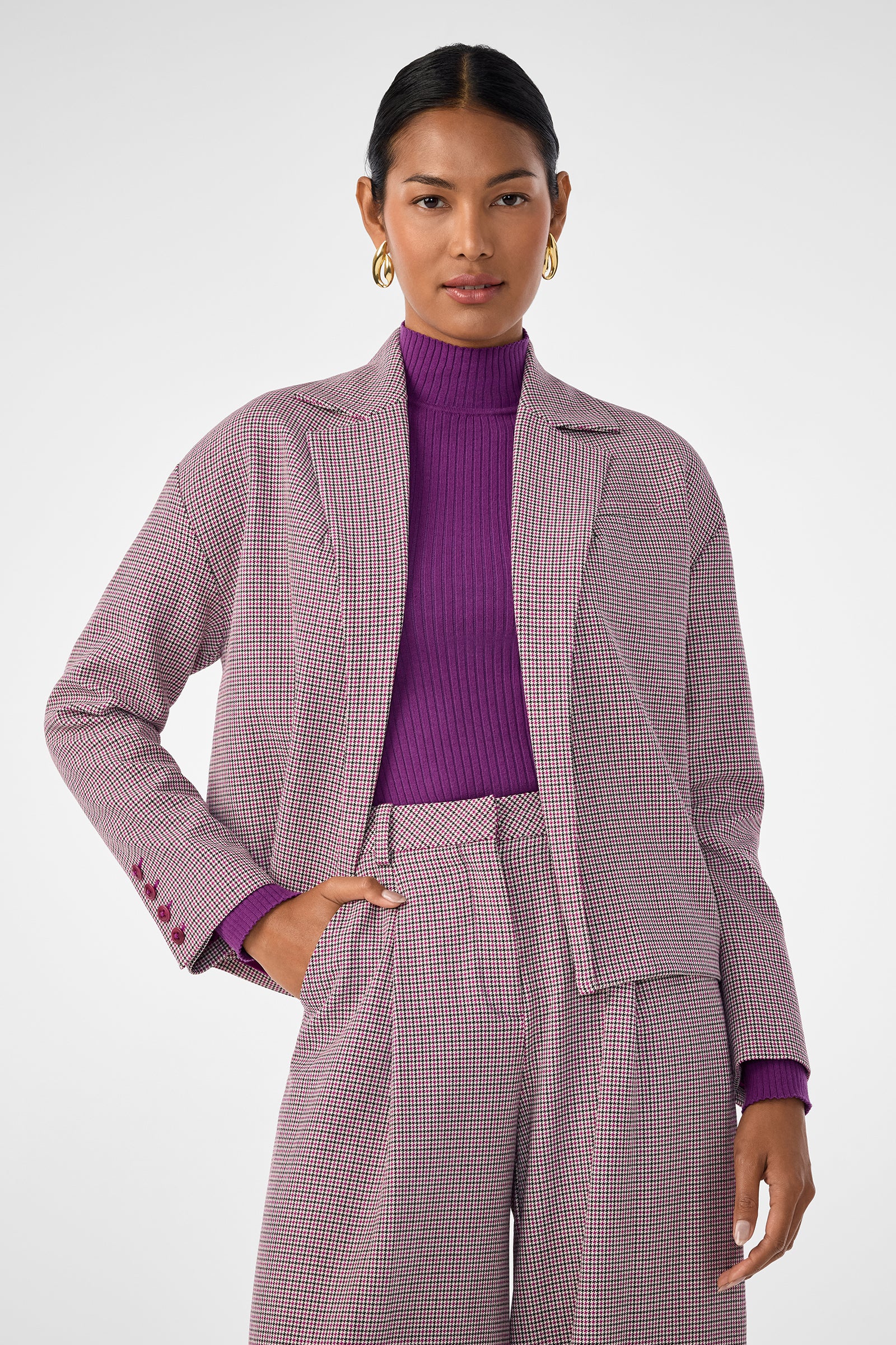 A woman with dark hair in a low bun wears gold hoops, a purple ribbed turtleneck, wide-leg plaid trousers, and the Ayla Jacket—a relaxed blazer—while standing against a light gray background.