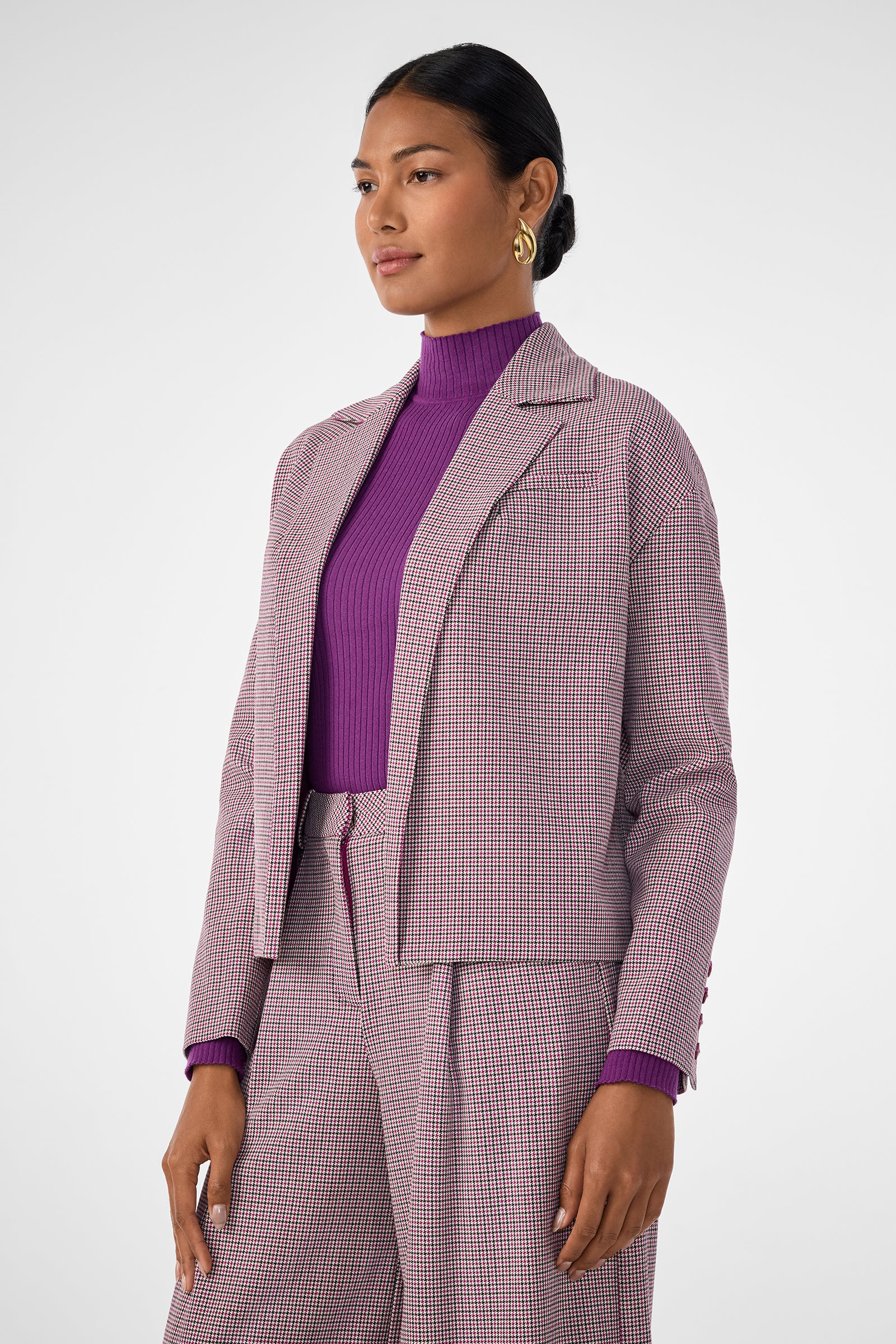 A woman stands against a plain background wearing the Ayla Jacket with matching wide-leg trousers and a purple ribbed turtleneck. Her hair is tied back, and she accessorizes with gold hoop earrings.