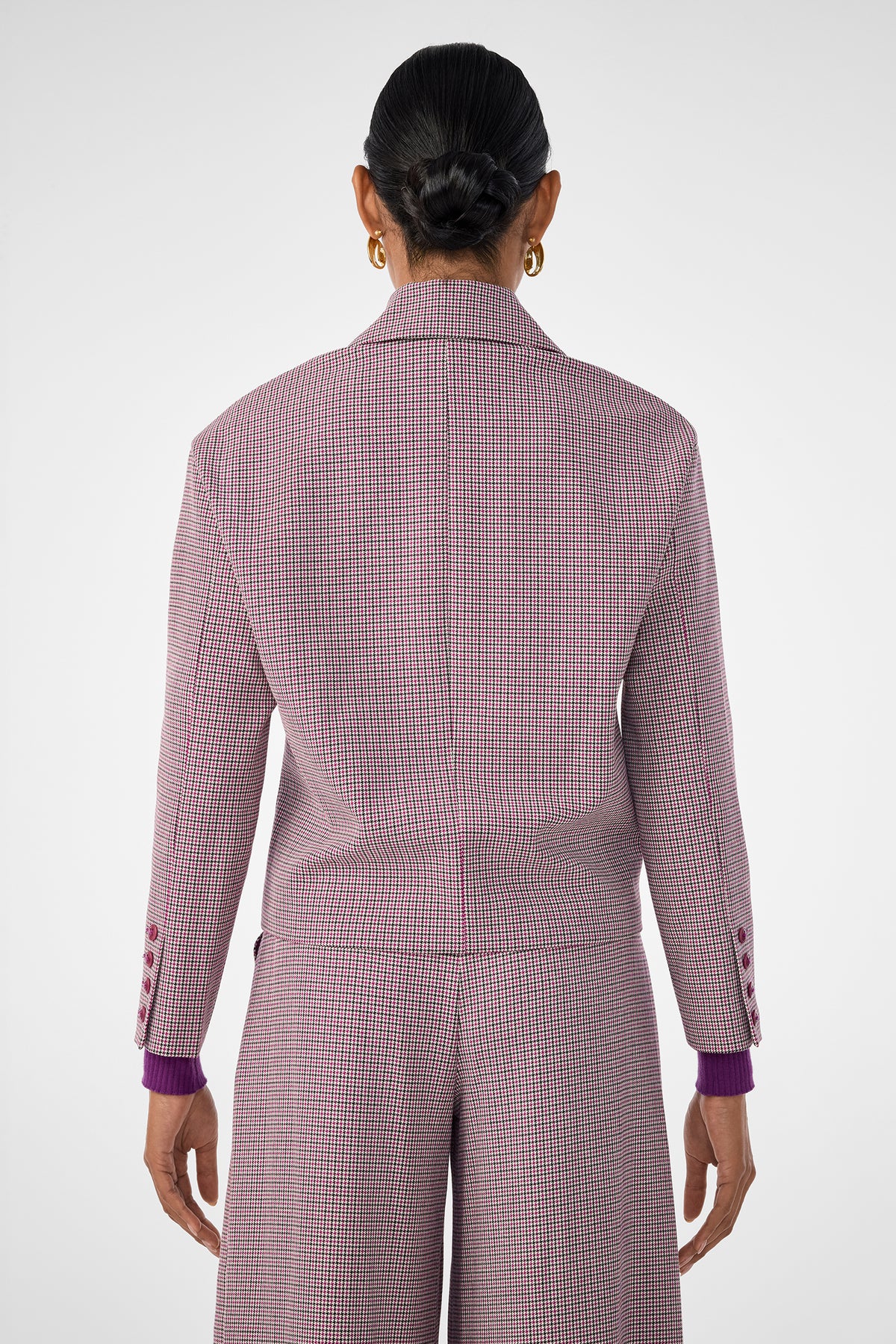 A person with dark hair in a neat bun is shown from behind, wearing the pink and white checkered Ayla Jacket with matching wide-leg pants and gold hoop earrings against a plain, light-colored background.