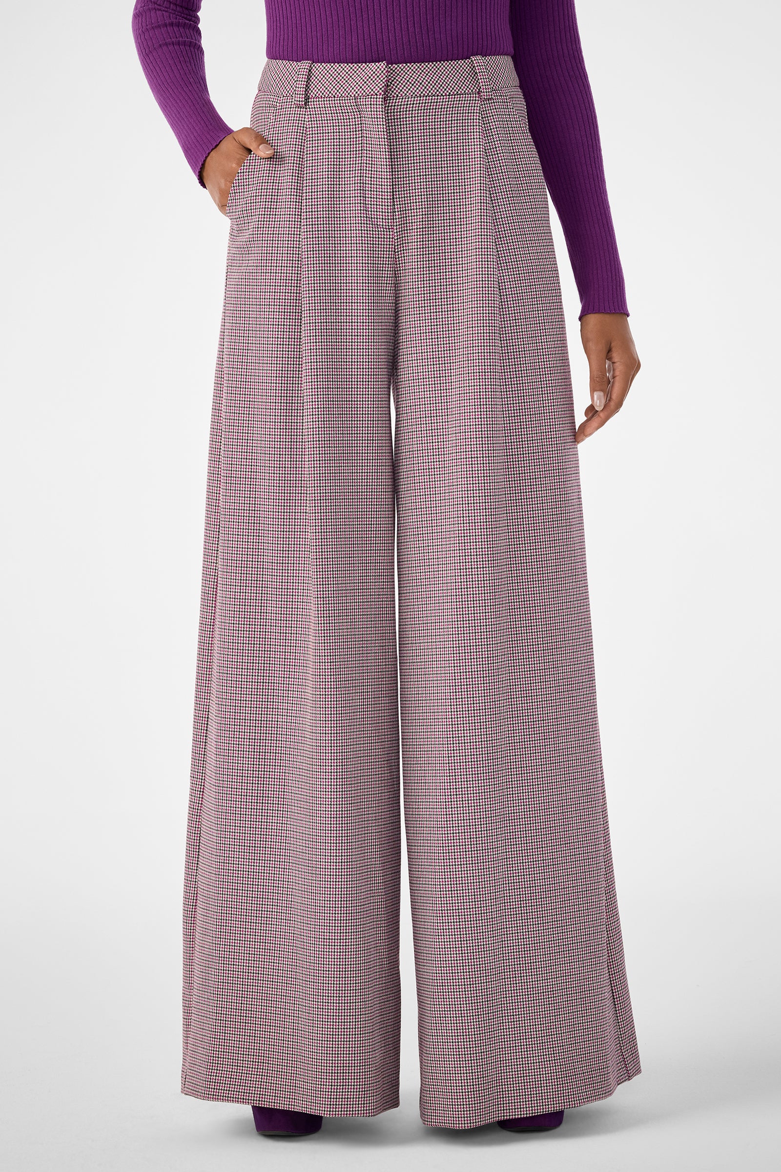 A person wears the Solani Pant, featuring high-waisted, wide-leg plaid in pink and grey, paired with a purple ribbed long-sleeve top. One hand is in their pocket, standing against a plain background.