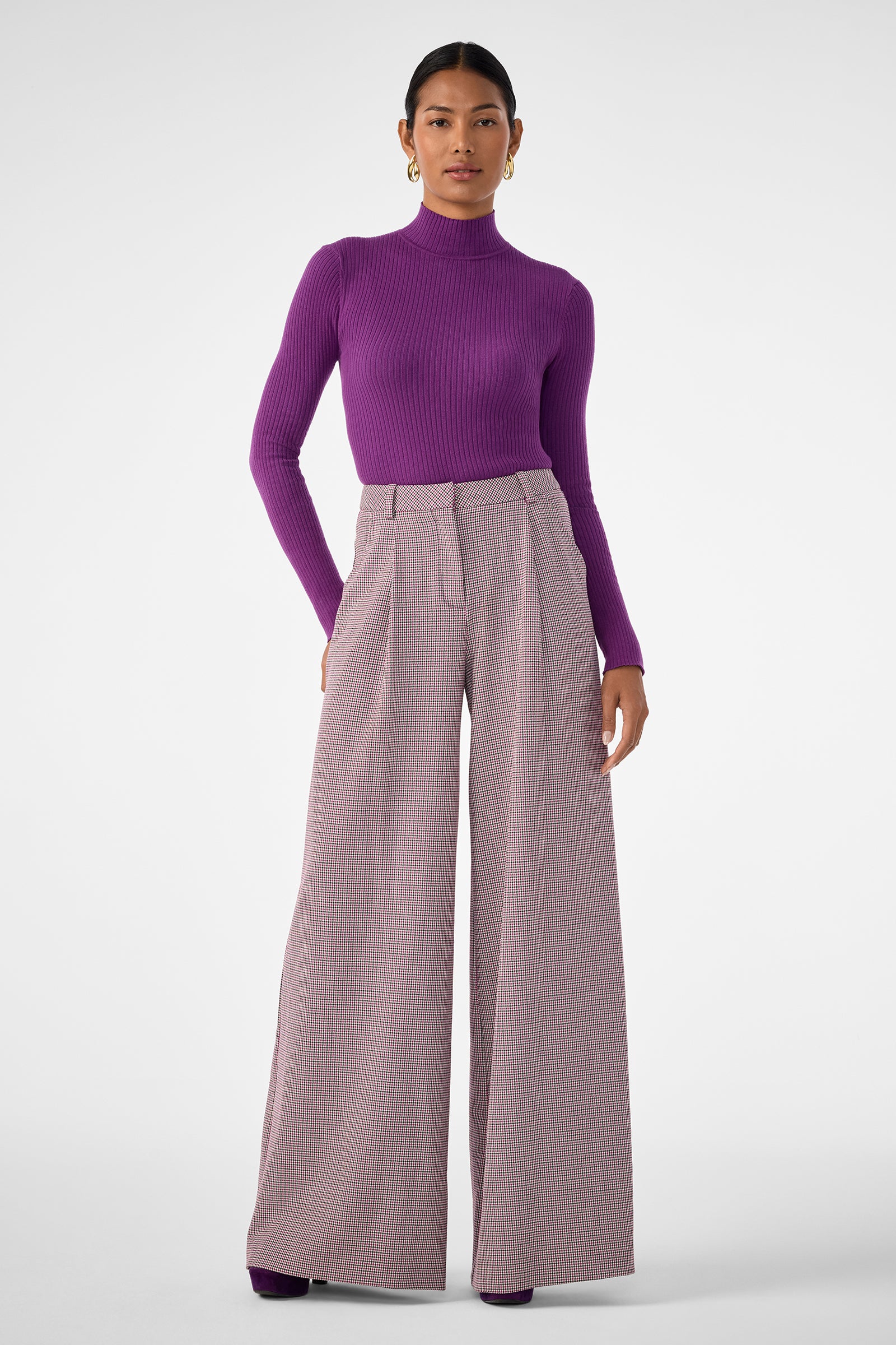 A woman stands against a plain background, confidently looking at the camera in high-waisted plaid Solani Pants, a fitted purple turtleneck sweater, and gold hoop earrings. Her hair is pulled back.