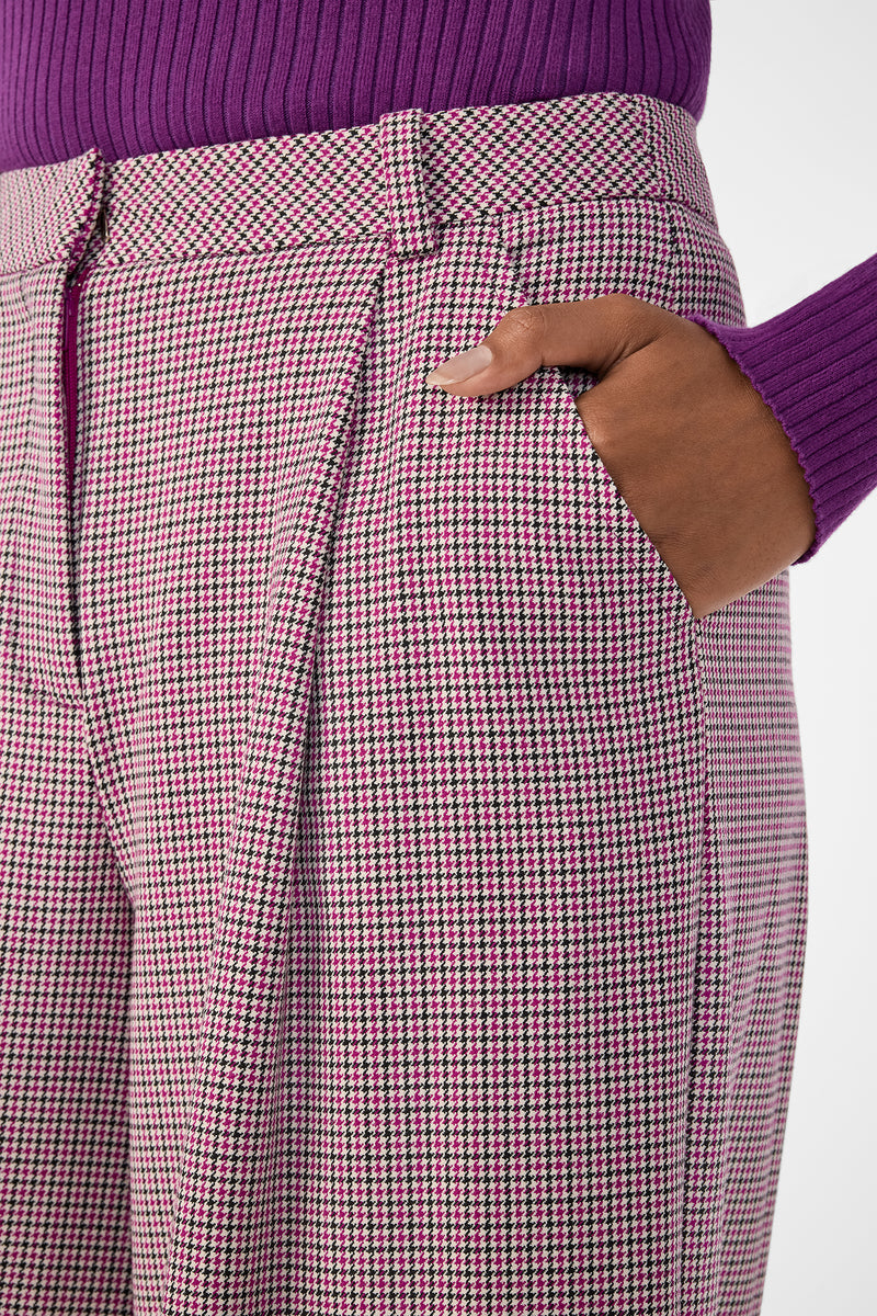 Someone stands with one hand in their pocket, wearing a purple ribbed sweater and high-waisted pink and black Solani Pants.