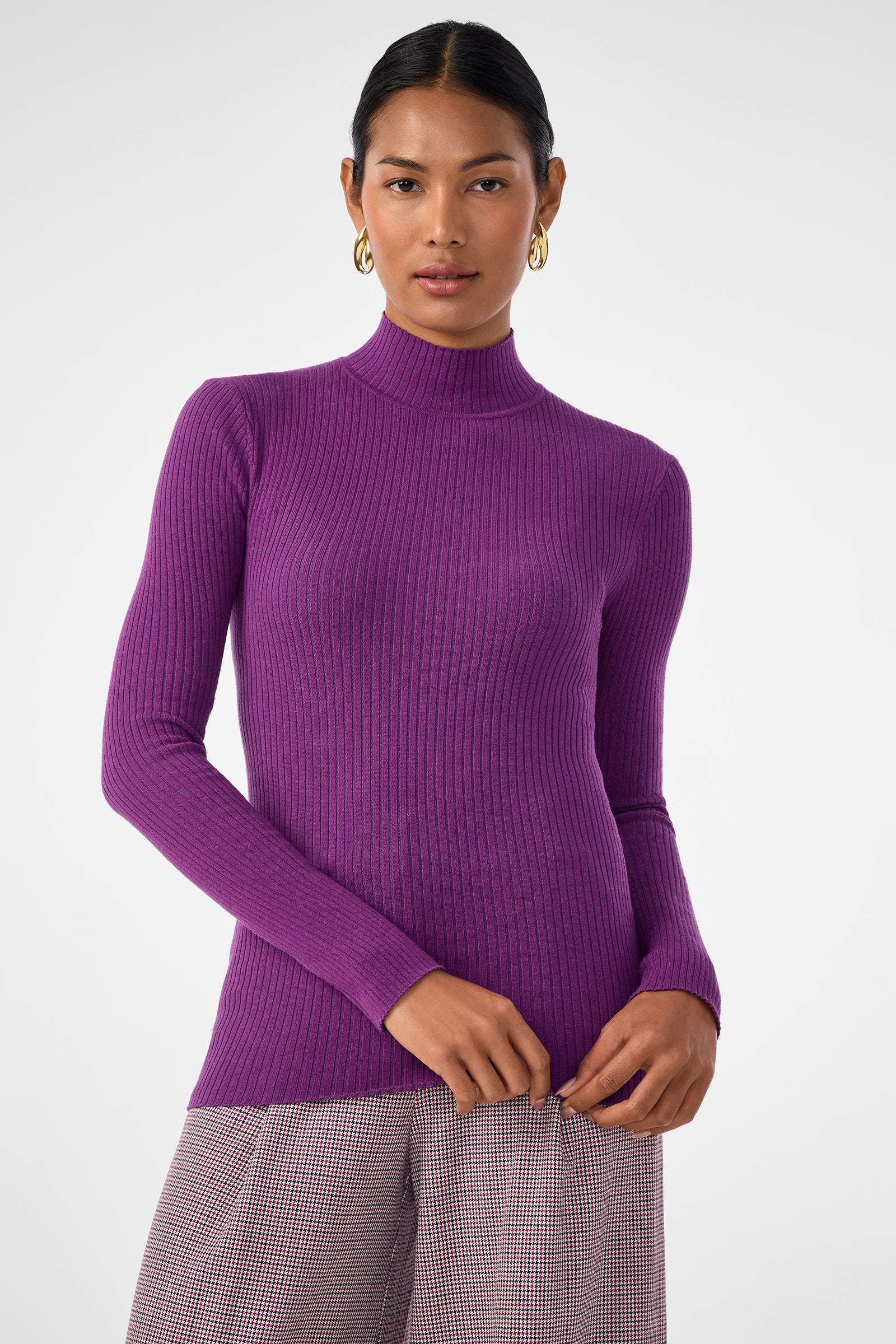 A woman stands against a plain background wearing the fitted, ribbed, long-sleeve purple Hempstead Sweater and checkered pants. She has gold hoop earrings and her dark hair is pulled back.