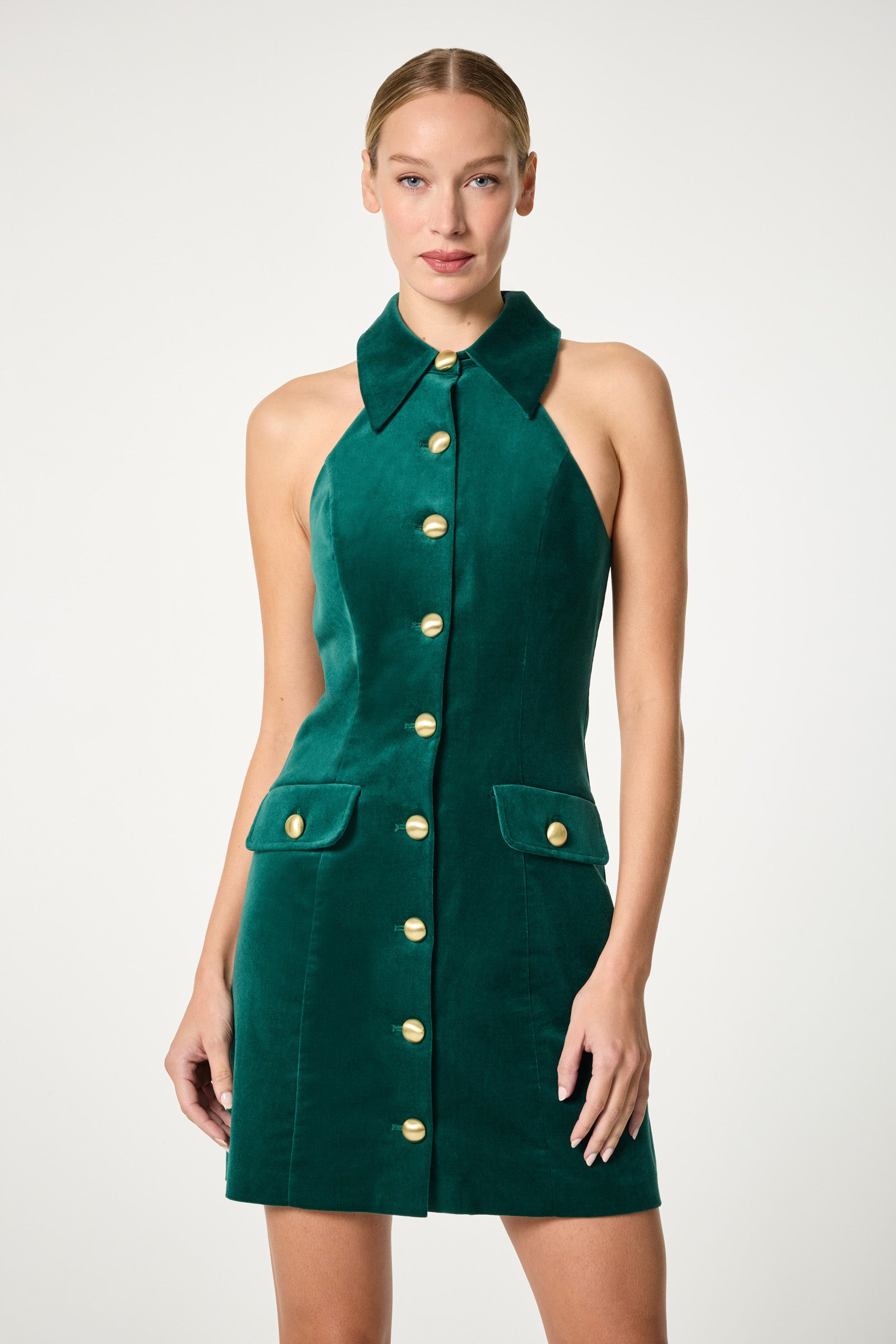 A woman wears the Safina Blazer Mini Dress, a sleeveless, emerald green velvet dress with a large collar, front gold buttons, and two front pockets, standing against a plain light background.
