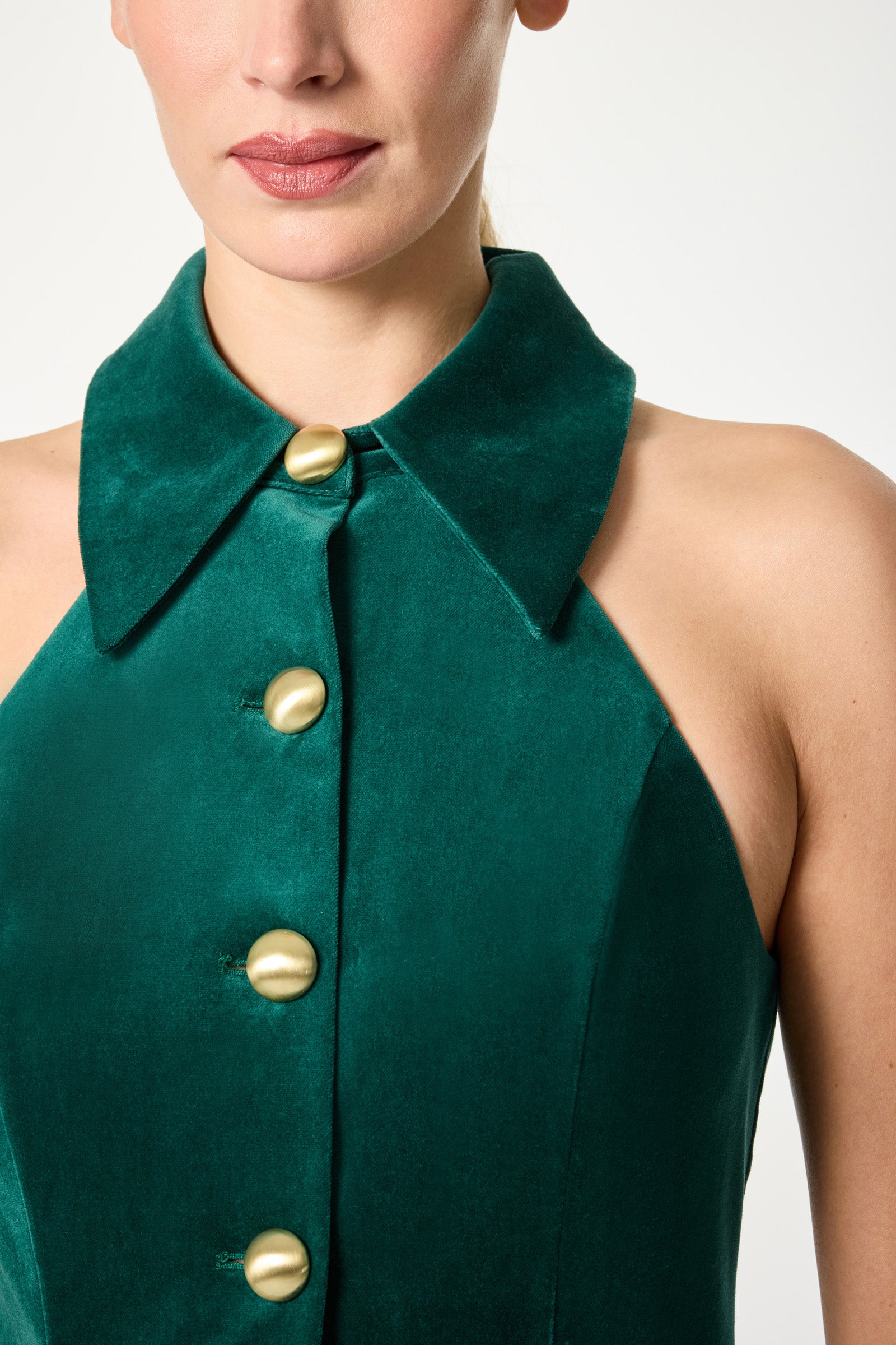 A woman wearing the Safina Blazer Mini Dress, a sleeveless emerald green velvet style with a wide collar and large gold front buttons. The image shows her from shoulders to just above the waist.