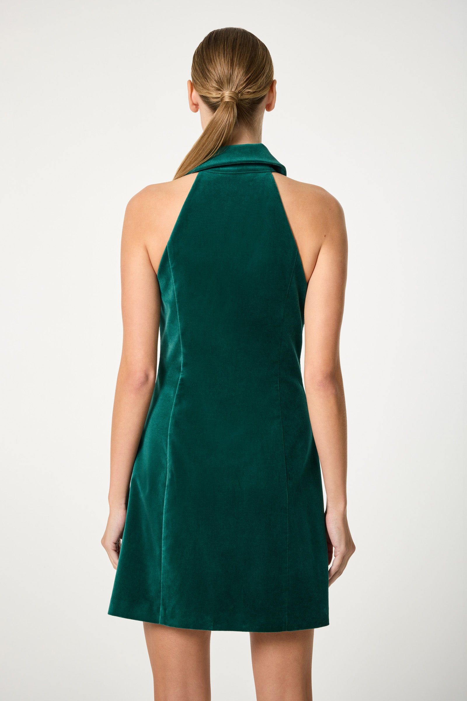 A woman with straight, light brown hair in a low ponytail is seen from behind wearing the Safina Blazer Mini Dress, a sleeveless green velvet halter style with a high collar, against a plain light background.
