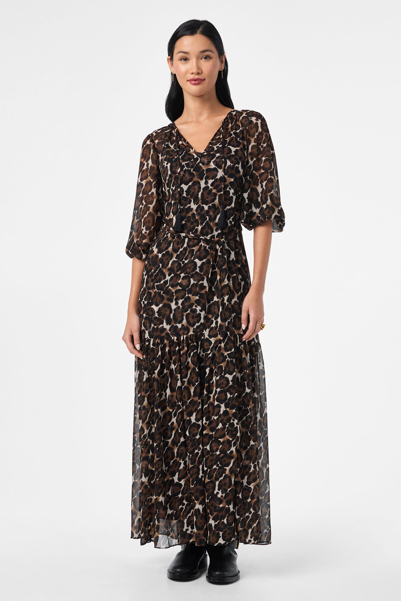 A woman stands against a white background wearing the Shalina Tie-Waist Maxi Dress, featuring a long, flowing leopard print design with three-quarter sleeves and paired with black shoes. She has straight dark hair and a neutral expression.