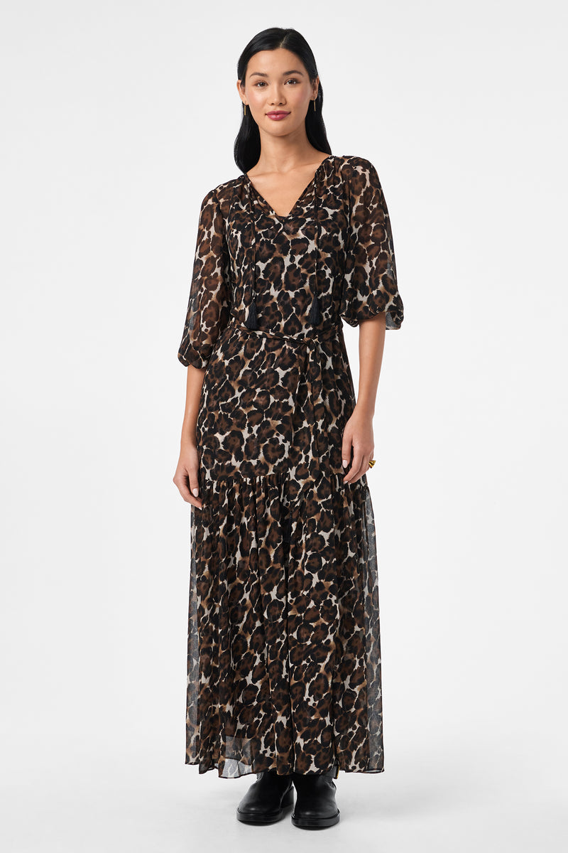 A woman stands against a white background wearing the Shalina Tie-Waist Maxi Dress, featuring a long, flowing leopard print design with three-quarter sleeves and paired with black shoes. She has straight dark hair and a neutral expression.