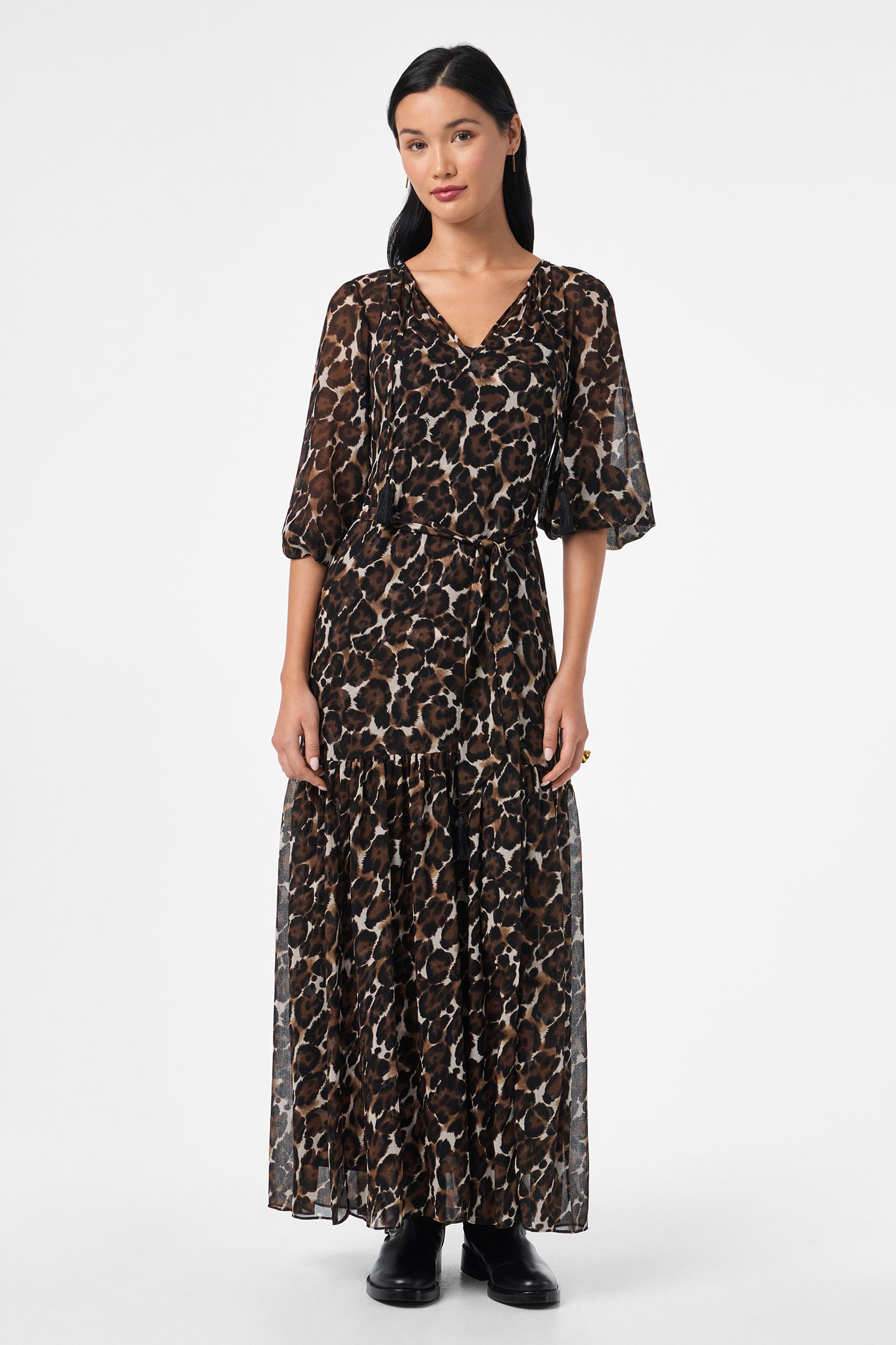 A woman faces forward in the Shalina Tie-Waist Maxi Dress, featuring a long, flowy leopard print with elbow-length sleeves and paired with black boots, set against a plain white background.