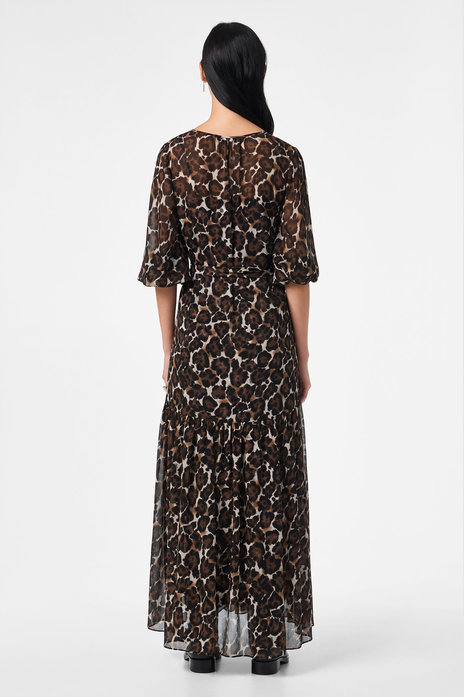 A person with long dark hair faces away, wearing the Shalina Tie-Waist Maxi Dress featuring a brown and black animal print, ankle length, and elbow-length sleeves against a plain white background.
