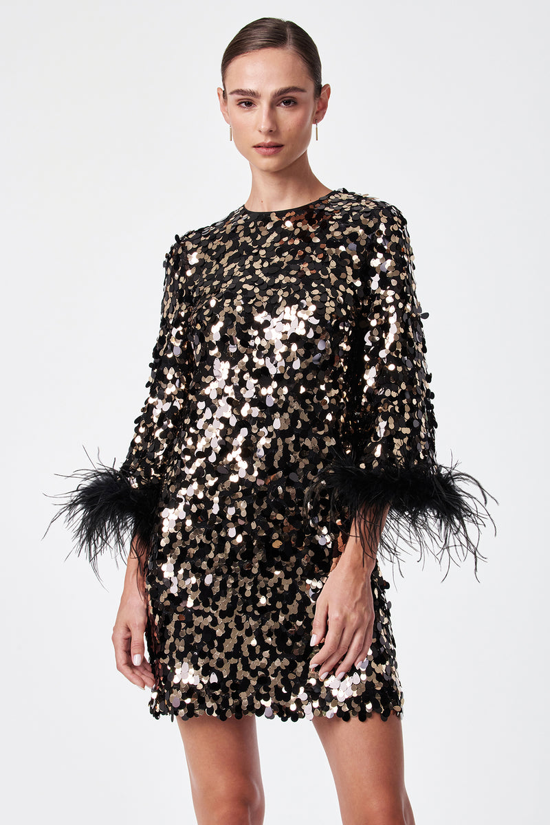 A woman models the Juliet Dress, a black and gold sequin outfit with black feather-accented sleeves, standing against a plain white background.