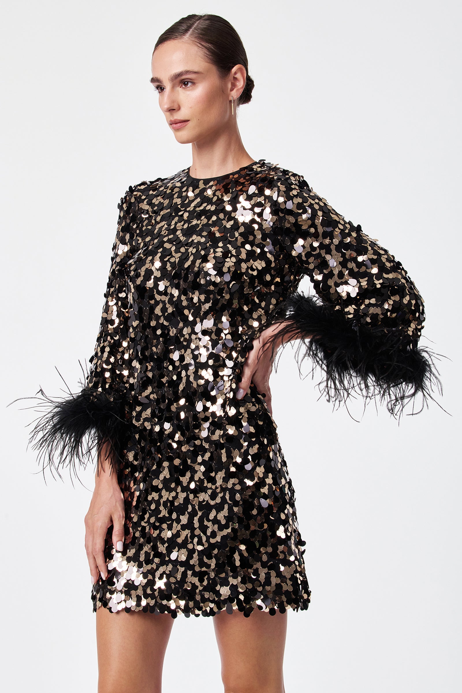 A woman models the Juliet Dress, a black long-sleeved mini dress with large gold sequins and black feathered cuffs, posing with one hand on her hip against a plain white background.